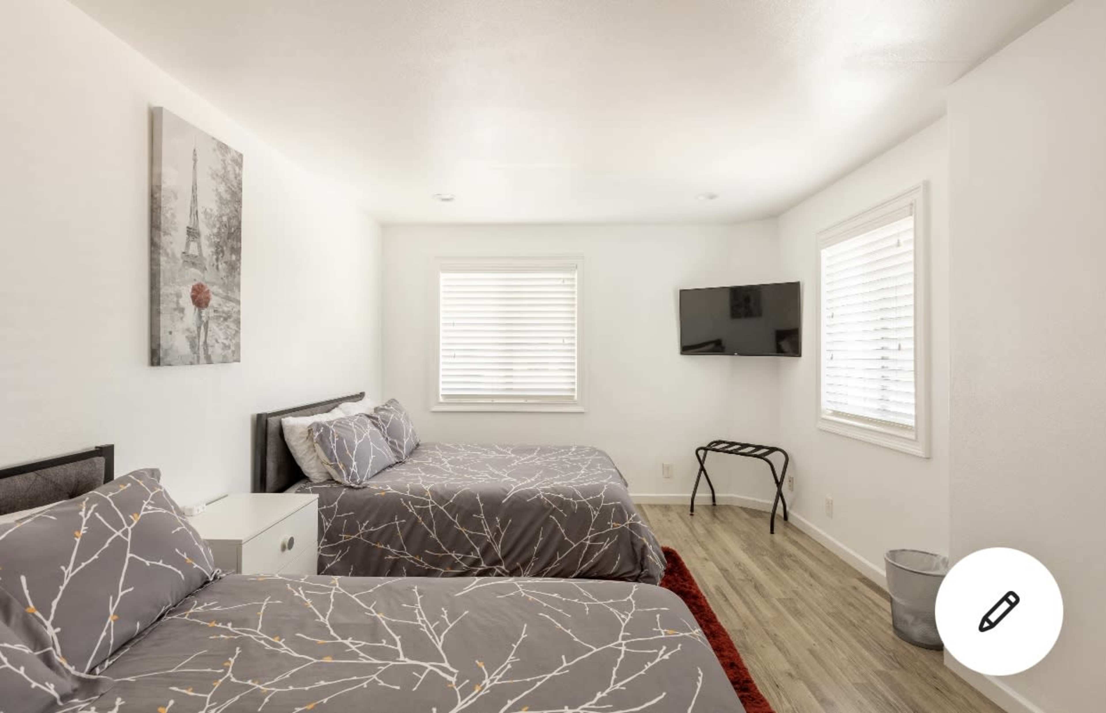The image shows a bright bedroom with two beds, a small nightstand, a television mounted on the wall, and a window with blinds.