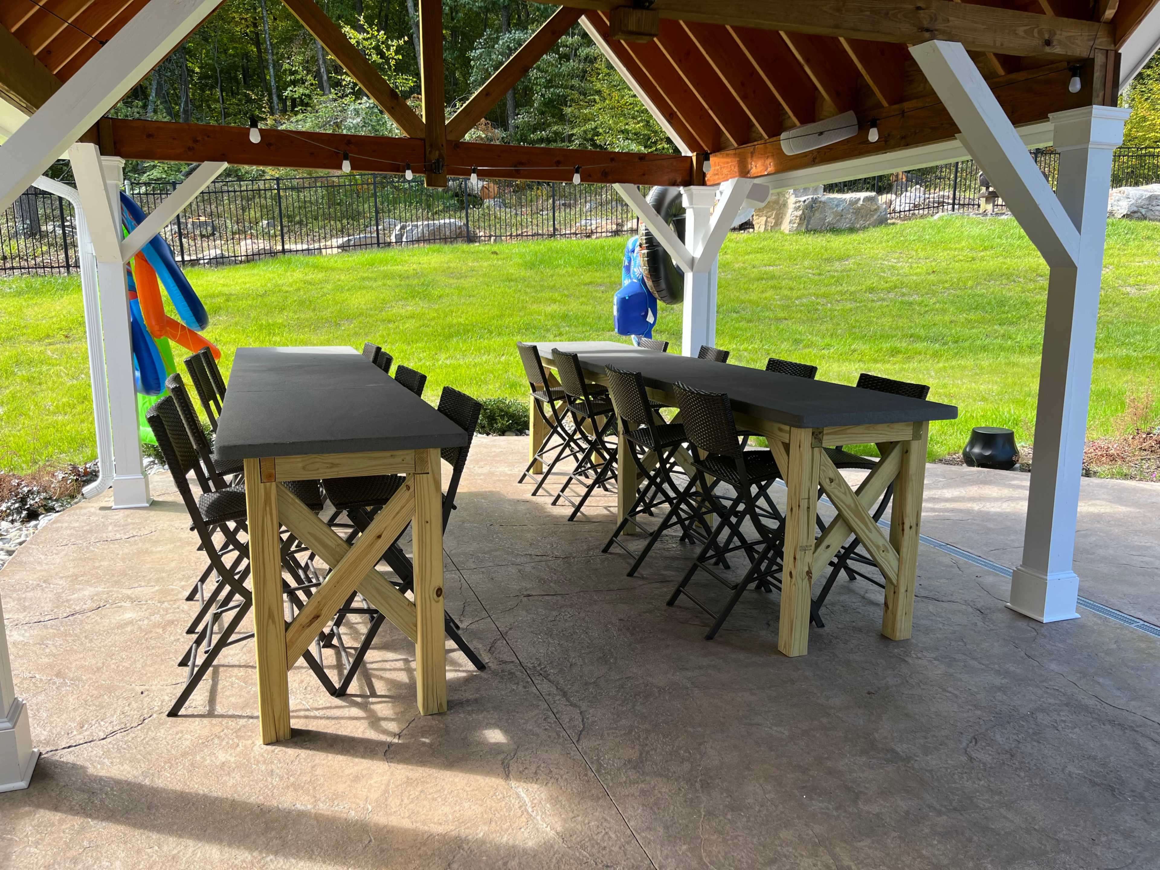 A covered outdoor pavilion features two long wooden tables with chairs arranged around them on a concrete floor.