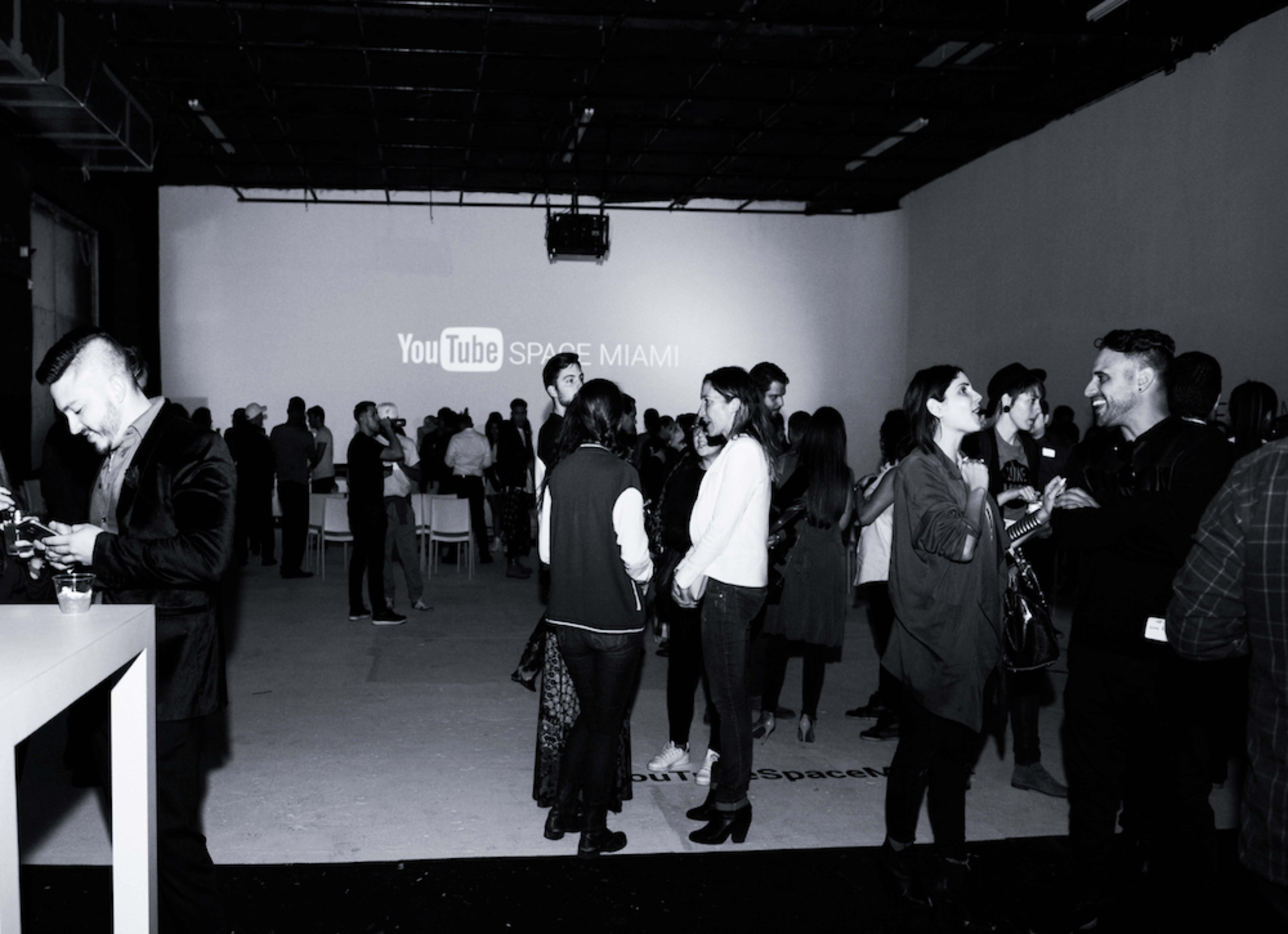 A group of people is gathered in a dimly lit space with a large screen displaying "YouTube SPACE MIAMI."