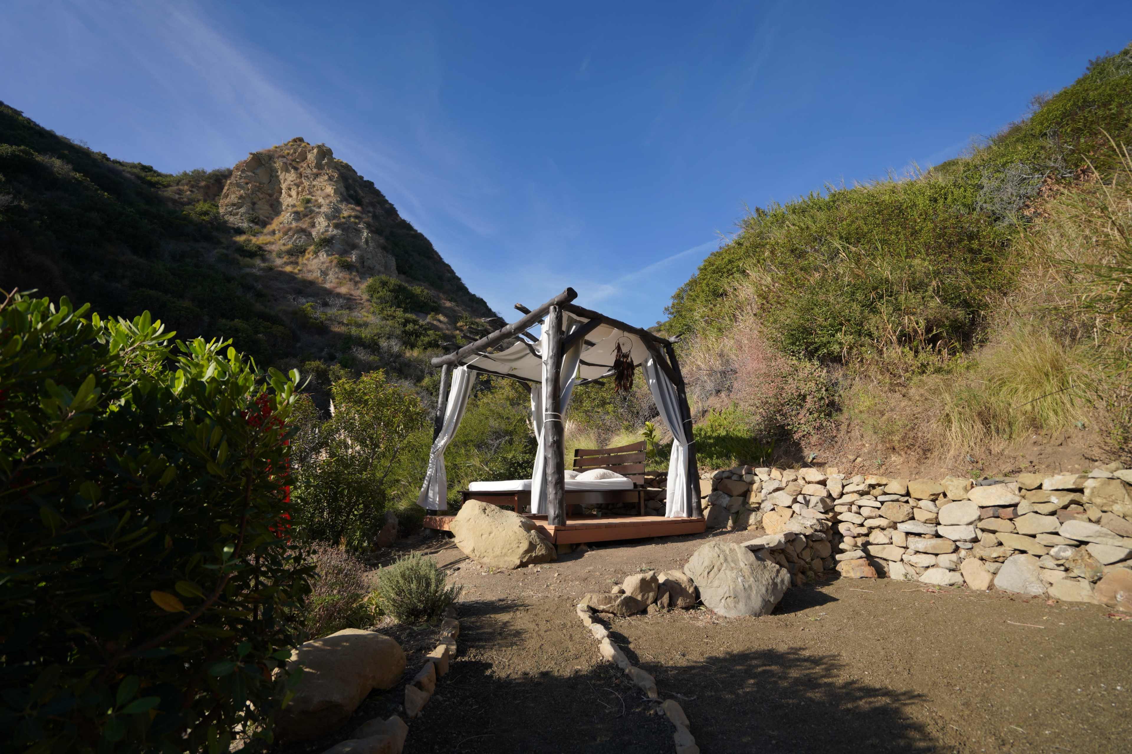 A wooden gazebo with draped fabric is set on a rocky pathway surrounded by greenery and hills.