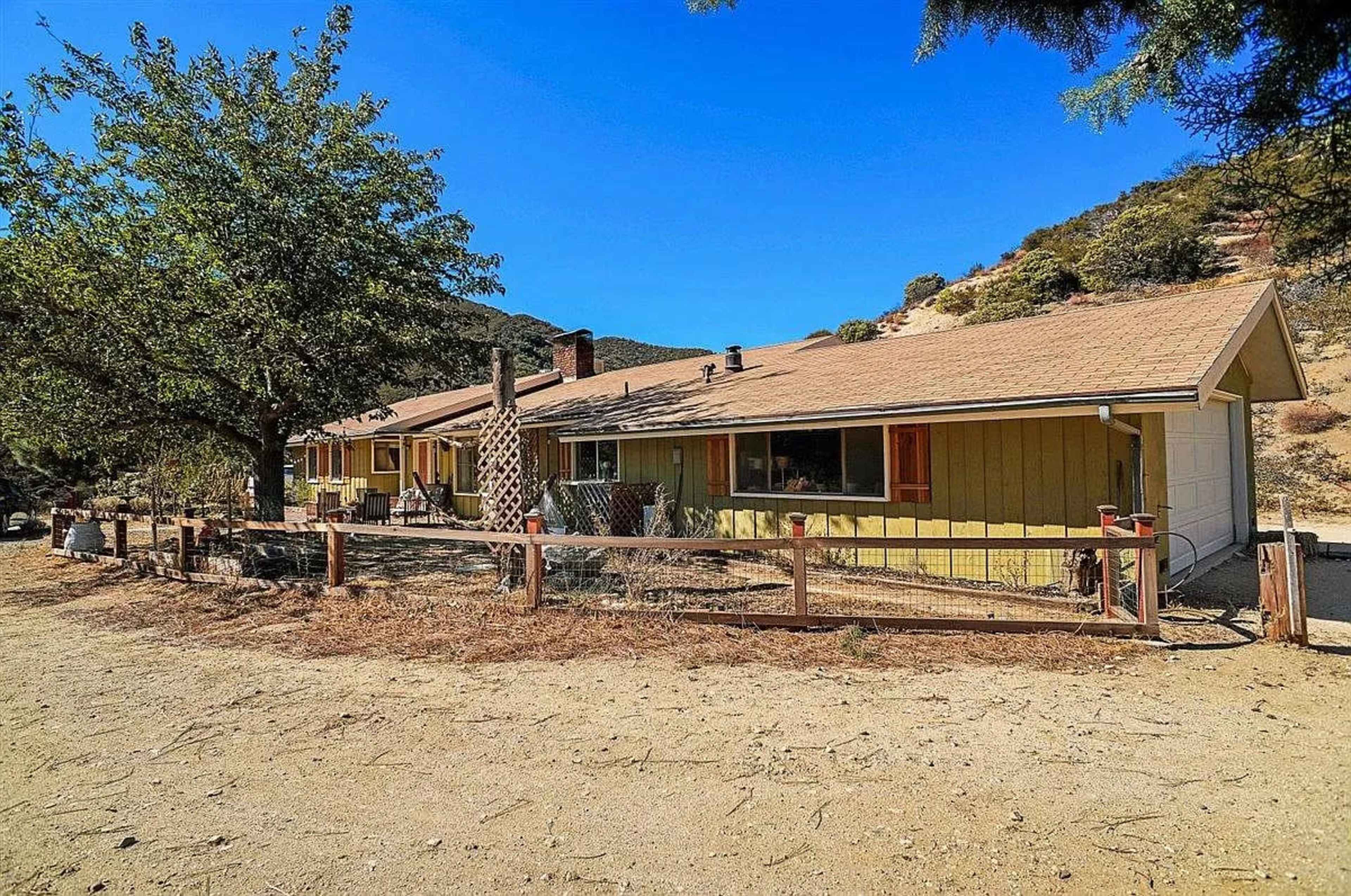 The image shows a yellow house with a brown roof, surrounded by a wooden fence and set against a mountainous background under clear blue skies.