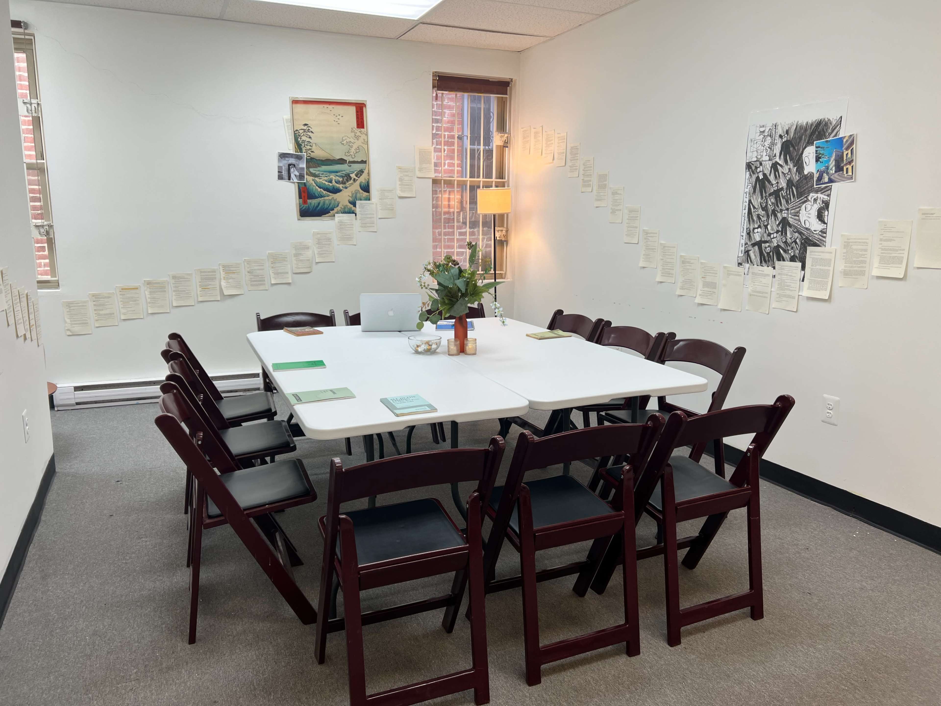 A conference room is arranged with a large table surrounded by chairs, and walls adorned with hanging papers and artwork.