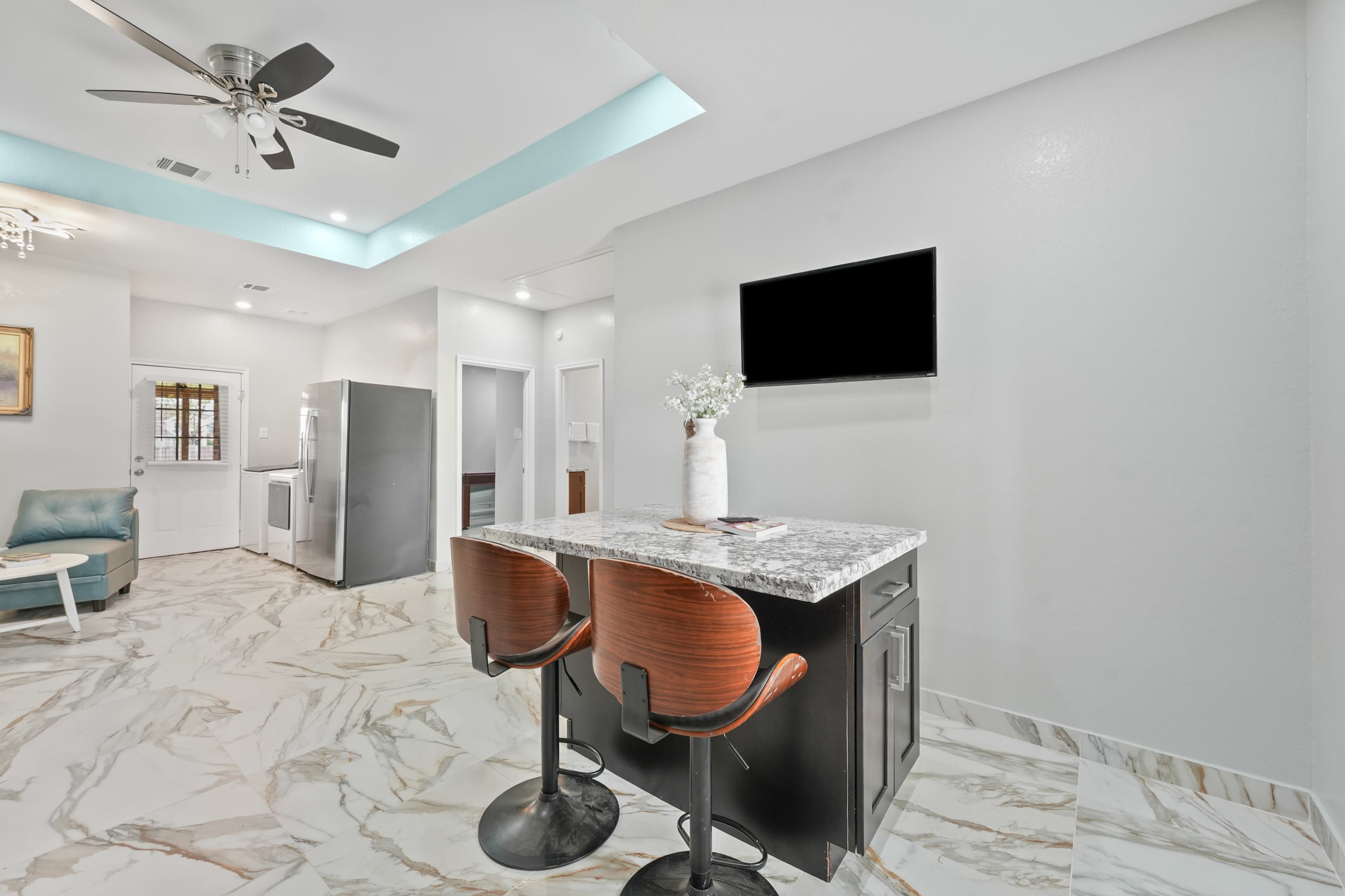The image shows a modern open-concept kitchen and living space featuring a granite countertop bar with two stools, stainless steel appliances, and light-colored walls.