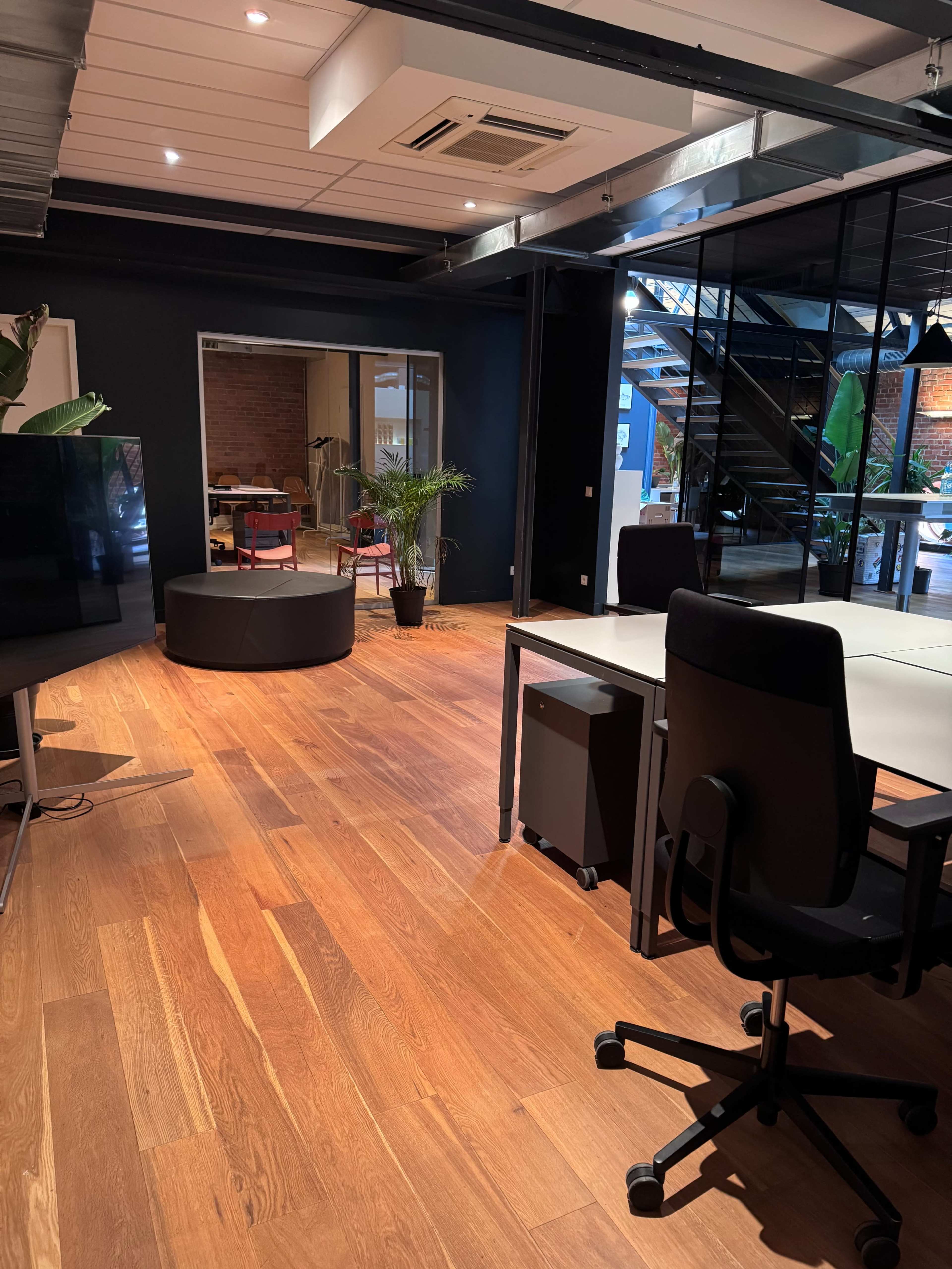 The image shows a modern office space with wooden flooring, several workstations, and a circular seating area.