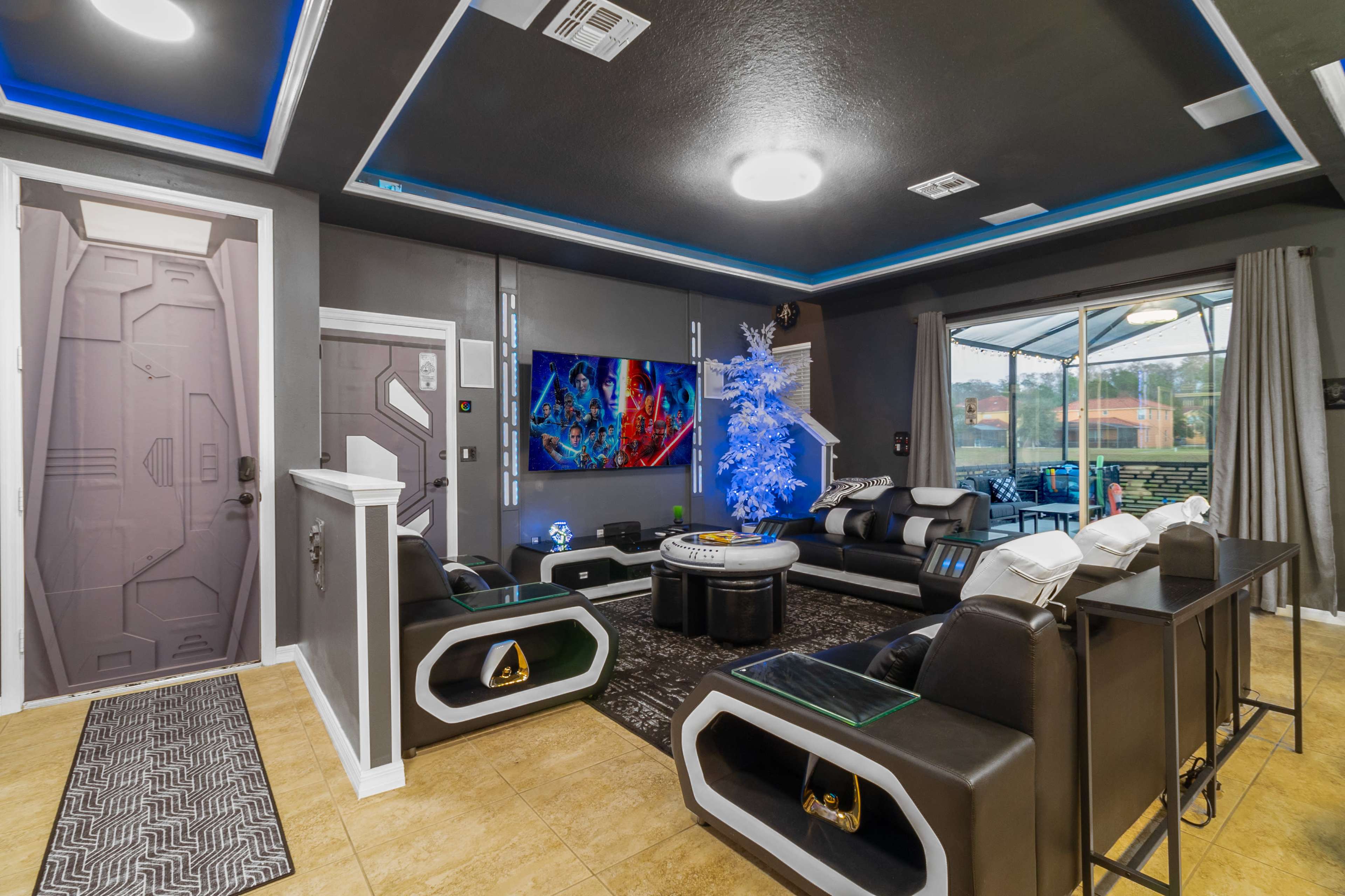 The image shows a modern living room with a futuristic design, featuring black furniture, a blue-lit decorative tree, and a large television displaying colorful artwork.