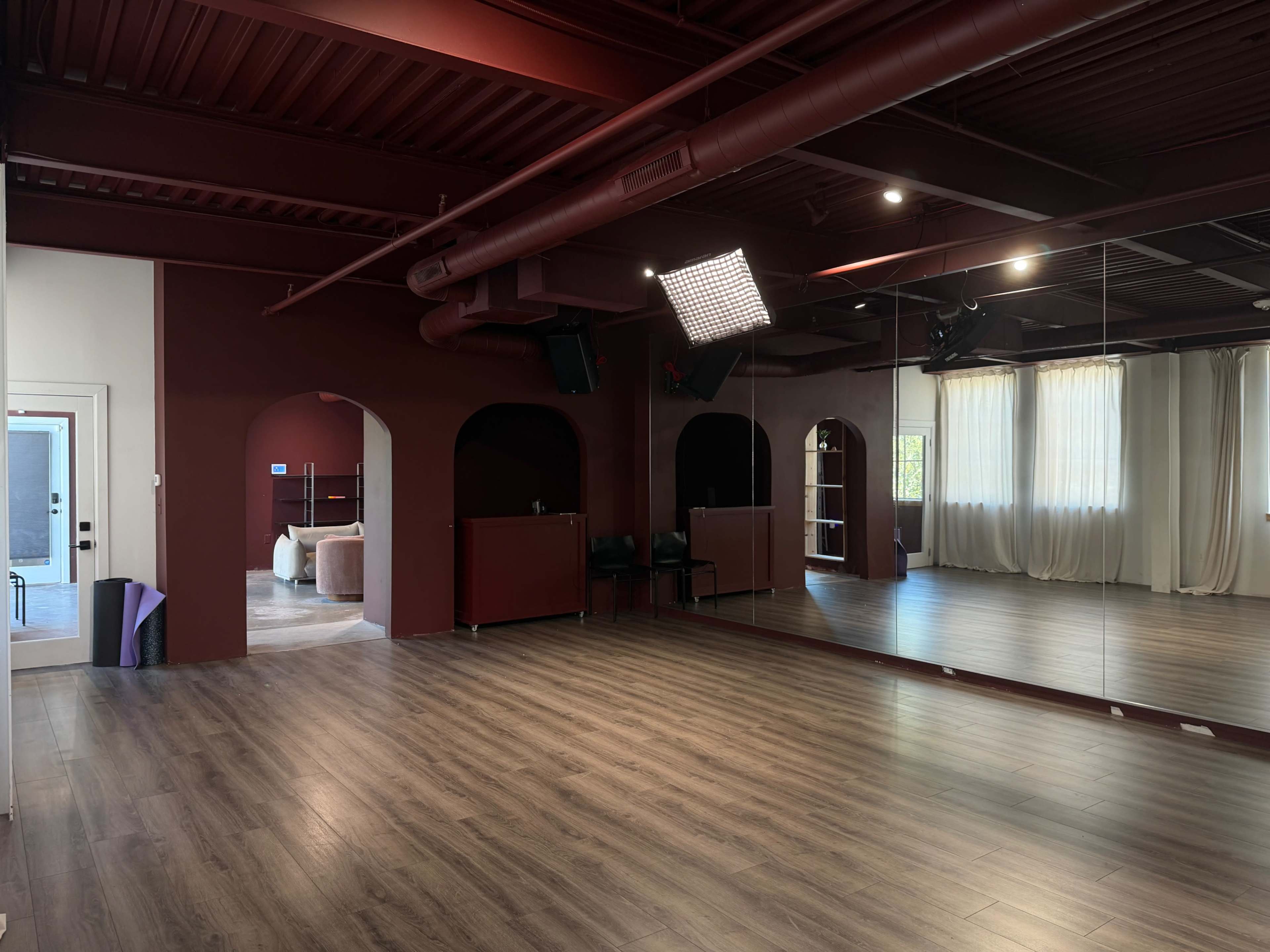 The image shows a spacious dance studio with maroon walls, wooden flooring, and a large mirror reflecting the room, which includes an arched doorway and soft white curtains letting in natural light.