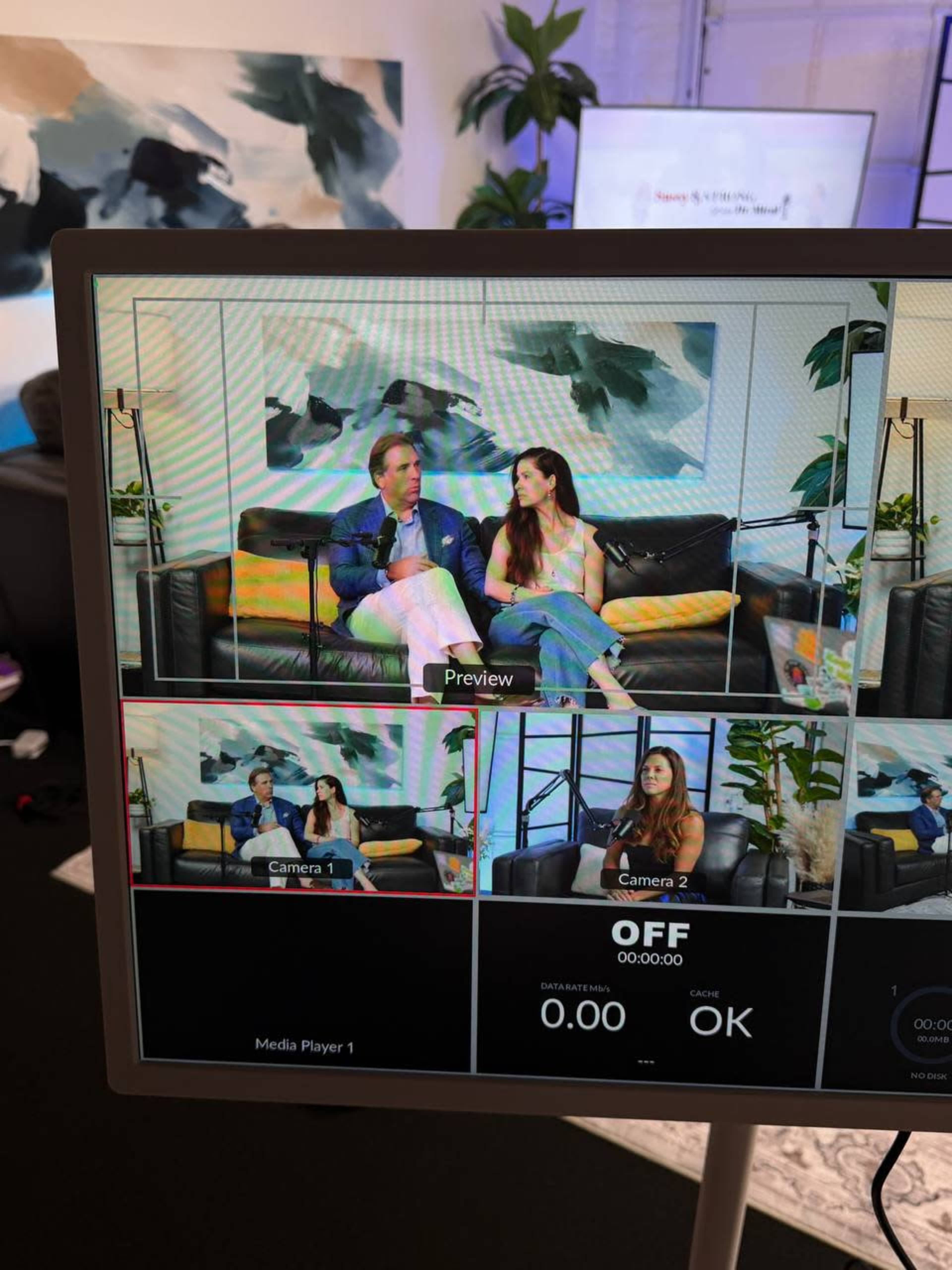 The image shows a video production setup with two people sitting on a couch, framed by multiple camera views on a monitor.