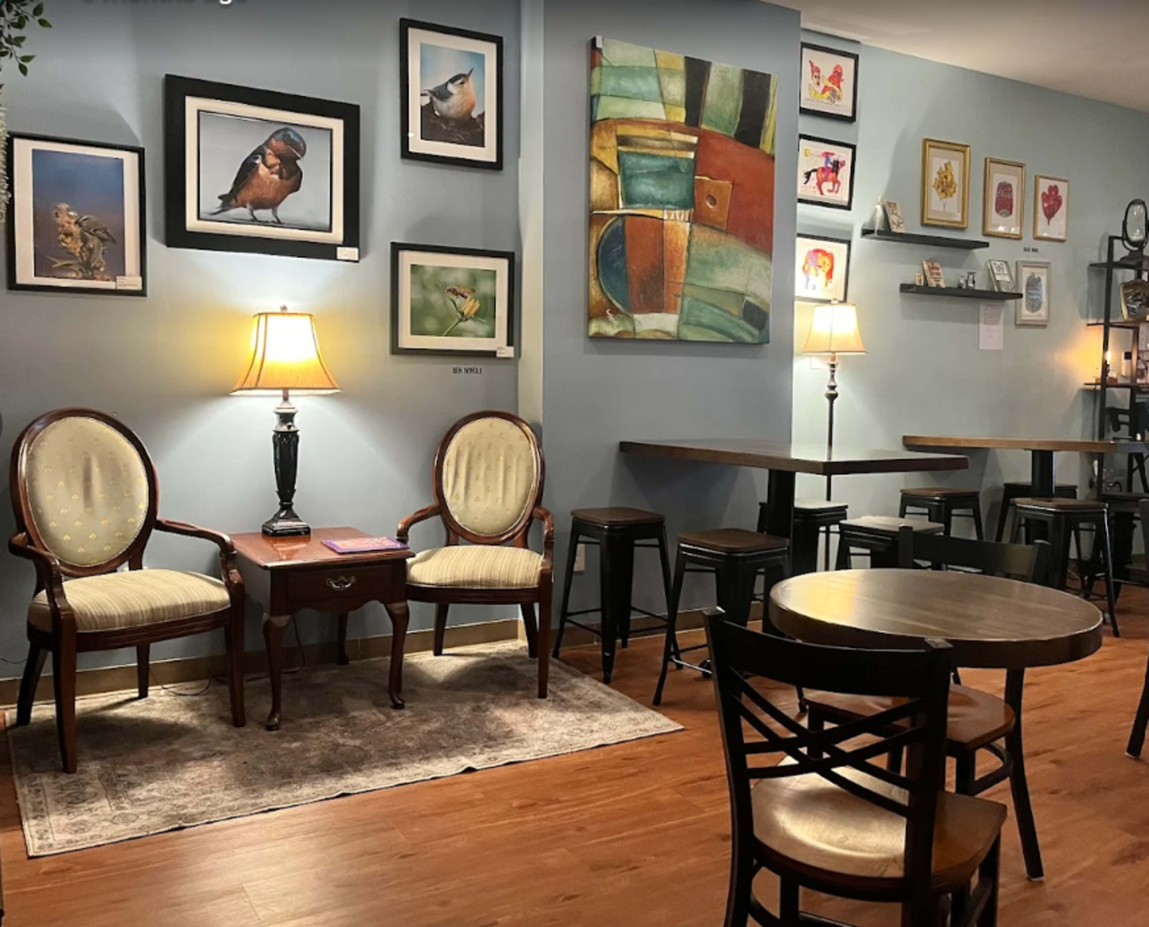 The image shows a cozy café interior featuring comfortable seating with two armchairs and a small table, framed artwork on the walls, and coffee tables arranged in a relaxed setting.