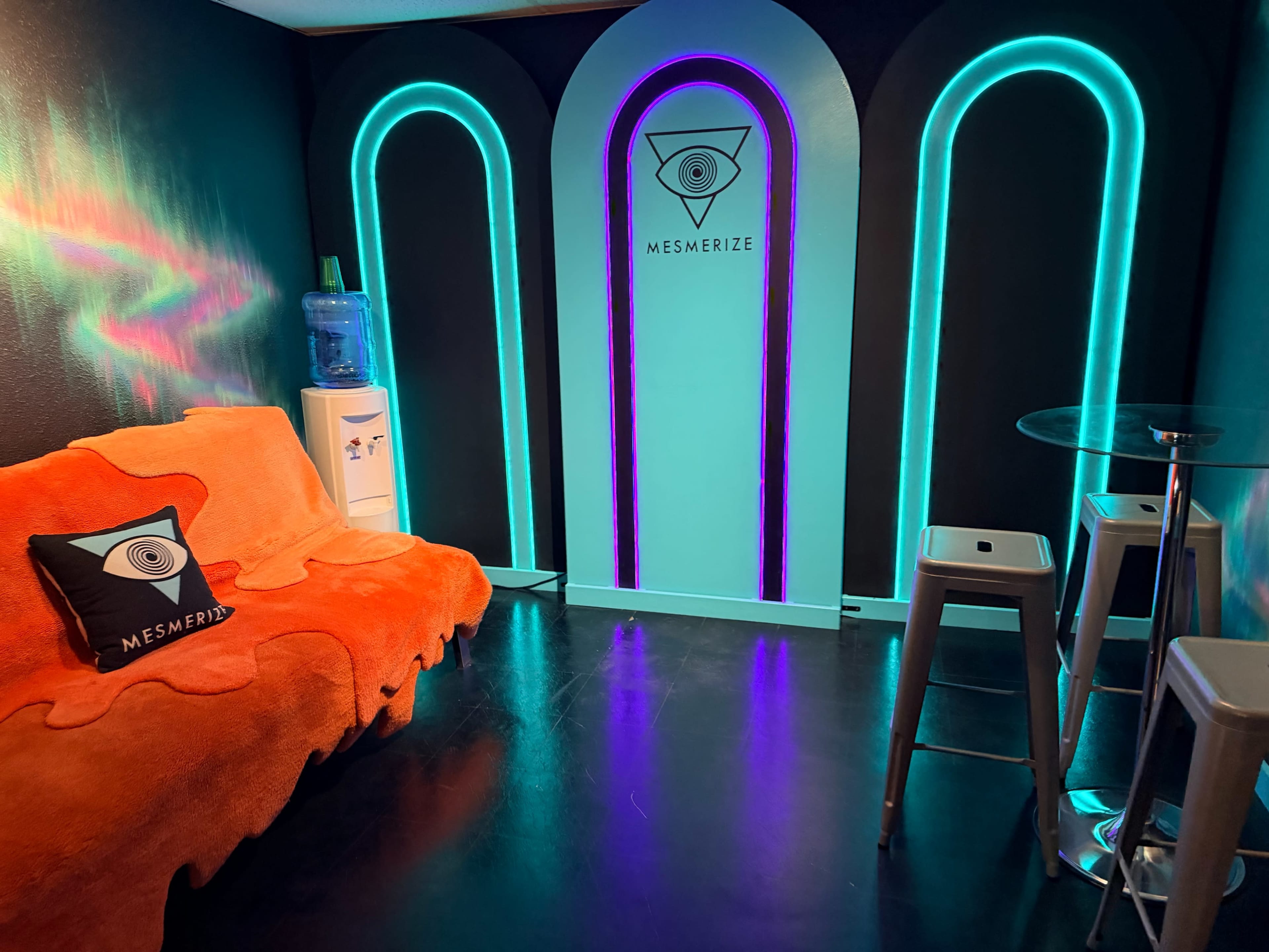The image shows a small lounge area featuring a bright orange couch, a glass table, a water cooler, and a wall design with neon blue and purple arches, along with the word "MESMERIZE" displayed on the wall.