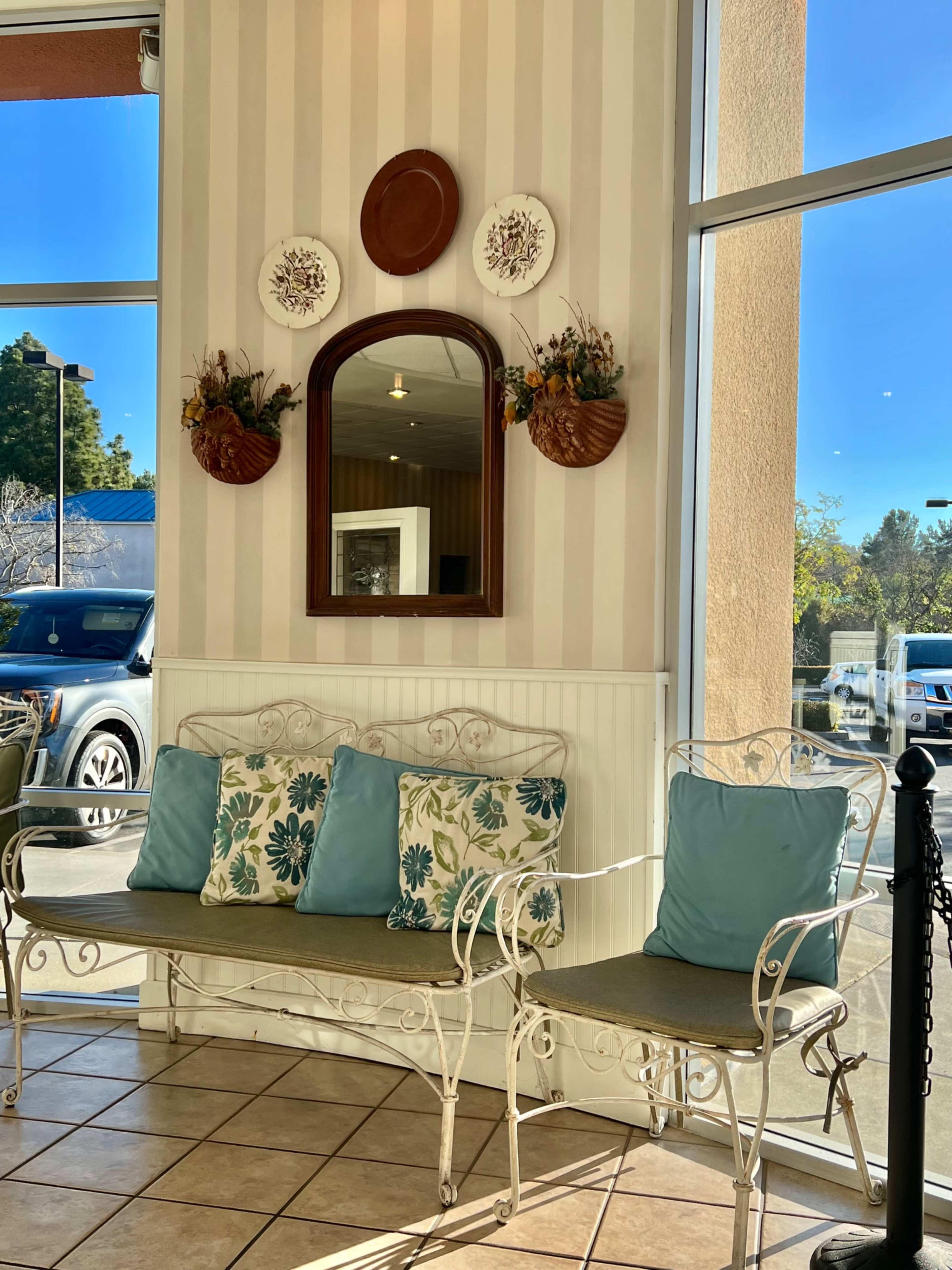 The image shows a seating area with a white metal bench and chairs adorned with blue and floral cushions, set against a wall featuring a mirror and decorative plates.
