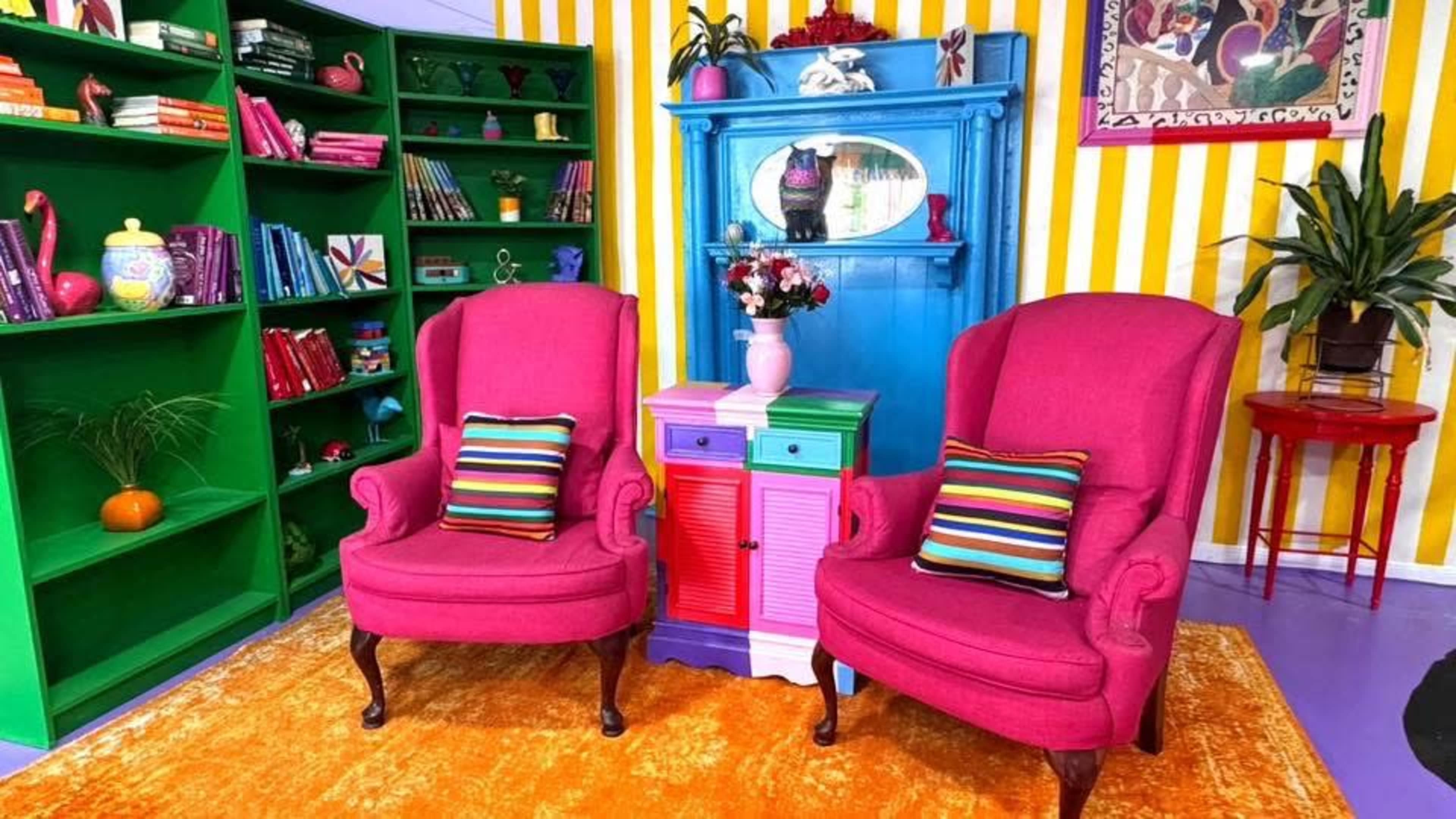 The image shows a vibrant room featuring two pink armchairs with colorful striped pillows, a multi-colored cabinet, a green bookshelf filled with books, and a patterned wall with yellow stripes.