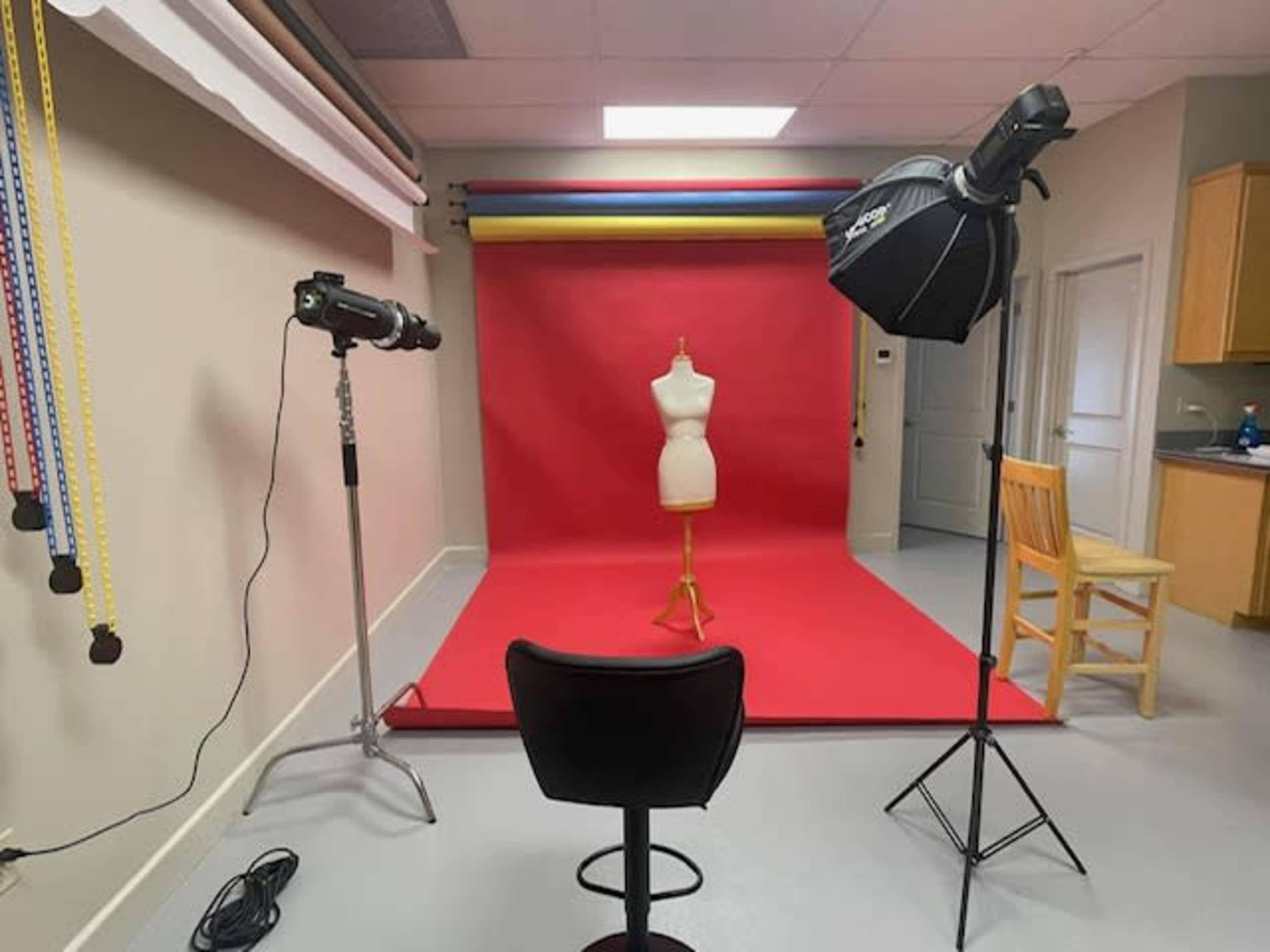 The image shows a photography studio with a red backdrop, a dress form on a stand, and two studio lights positioned for a photoshoot, along with a chair and a kitchenette in the background.