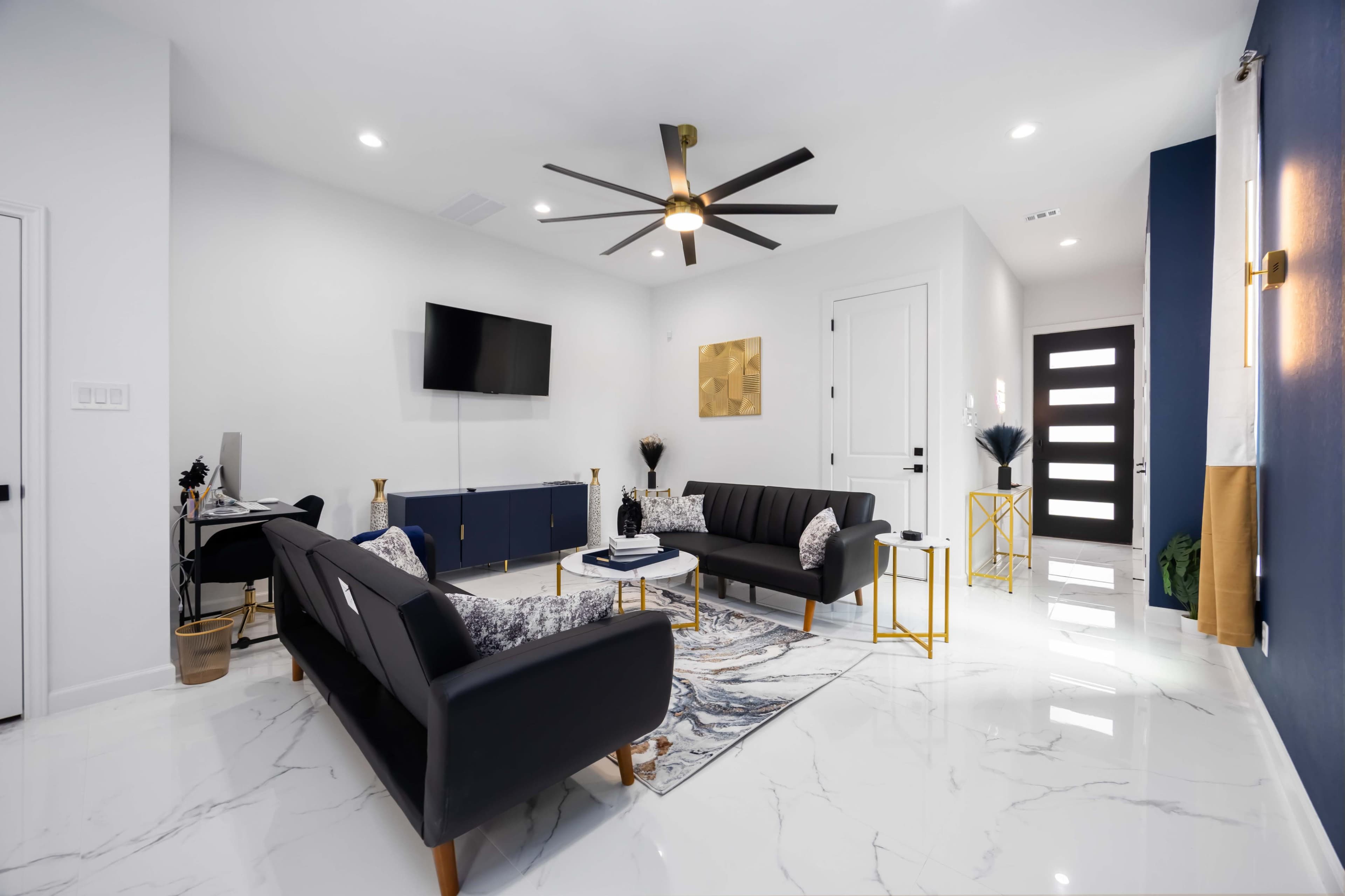 A modern living room features two black sofas, a marble coffee table, a large flat-screen TV mounted on a wall, and a front door with sleek glass panels.