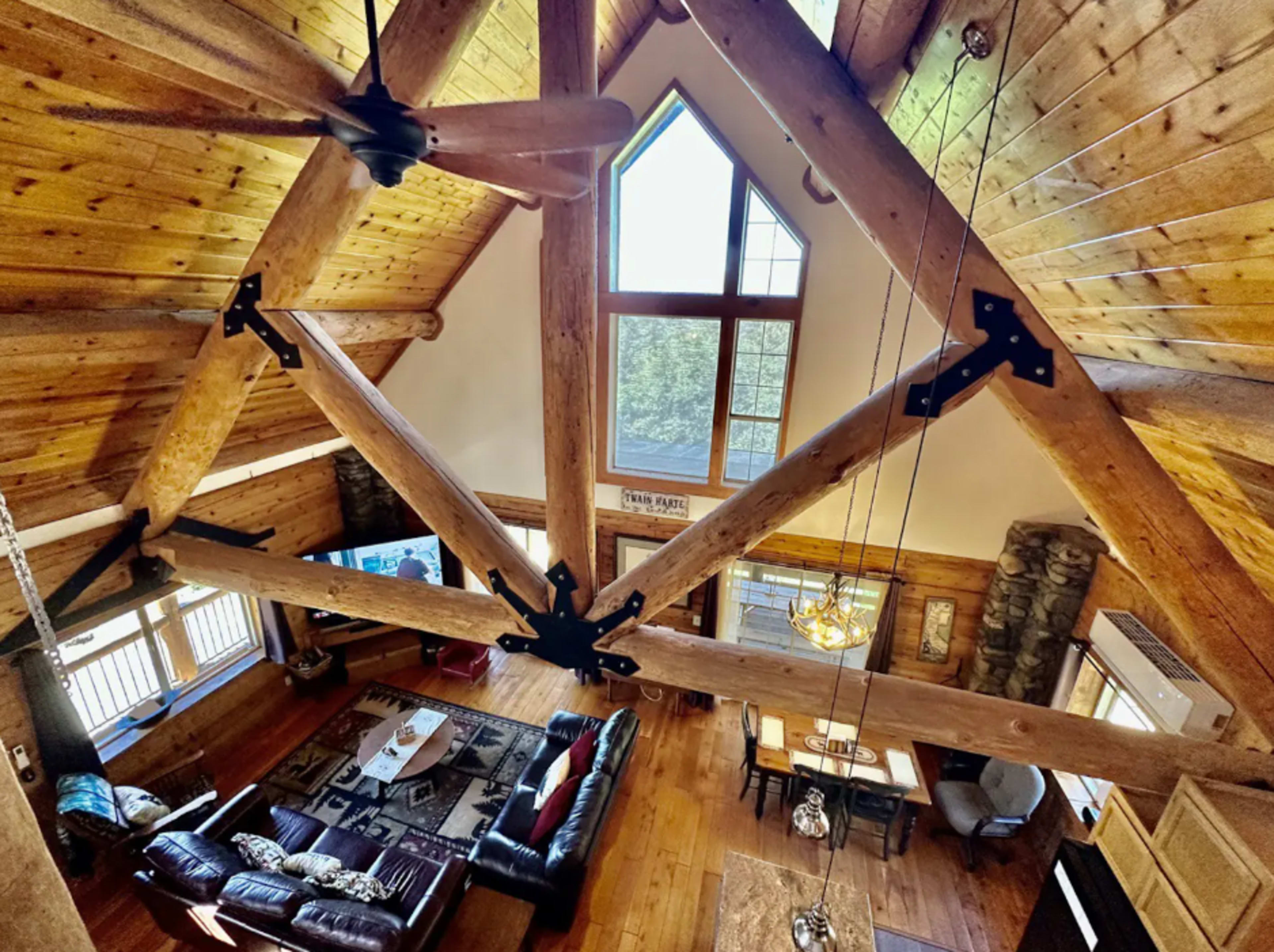 A spacious wooden lodge interior with exposed beams, a large window, and a living area furnished with a leather sofa and wooden decorations.