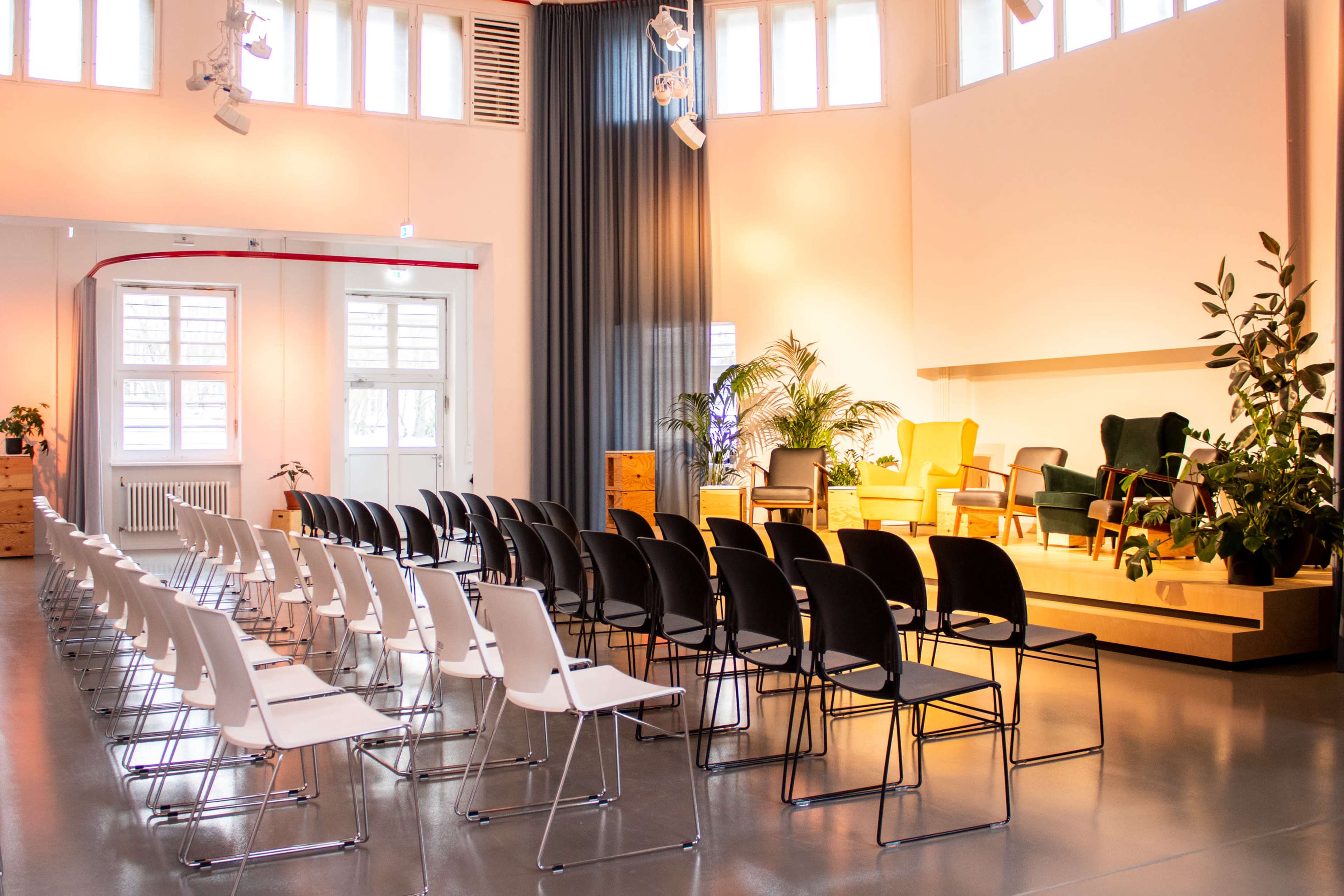 The image shows a seminar room arranged with rows of white and black chairs facing a small stage with a seating area surrounded by plants.
