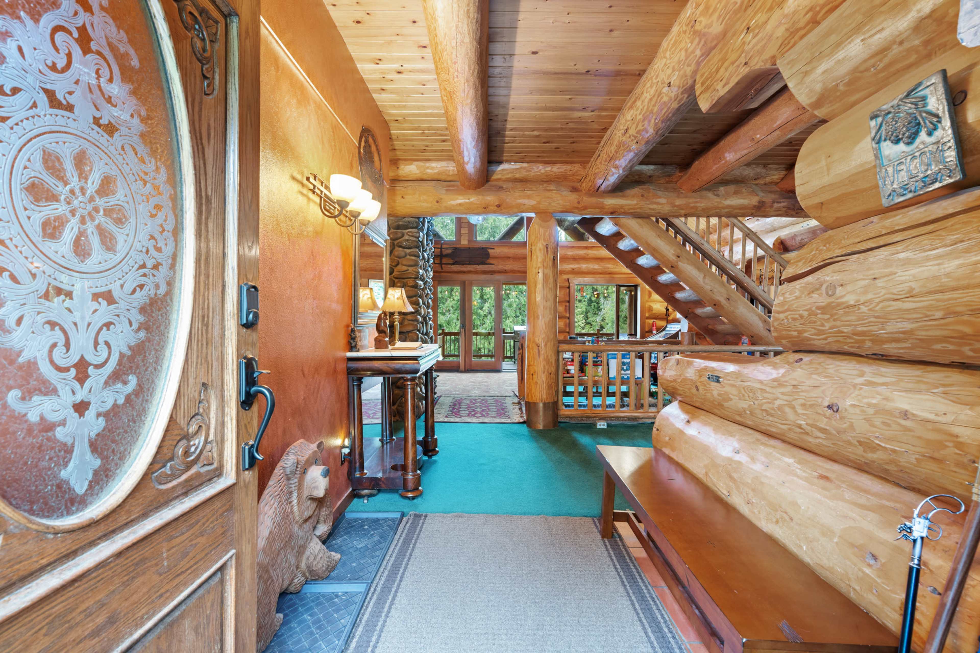 The image shows the interior of a log cabin entryway, featuring a wooden door, a staircase to the upper level, and a view of large windows overlooking a forest.