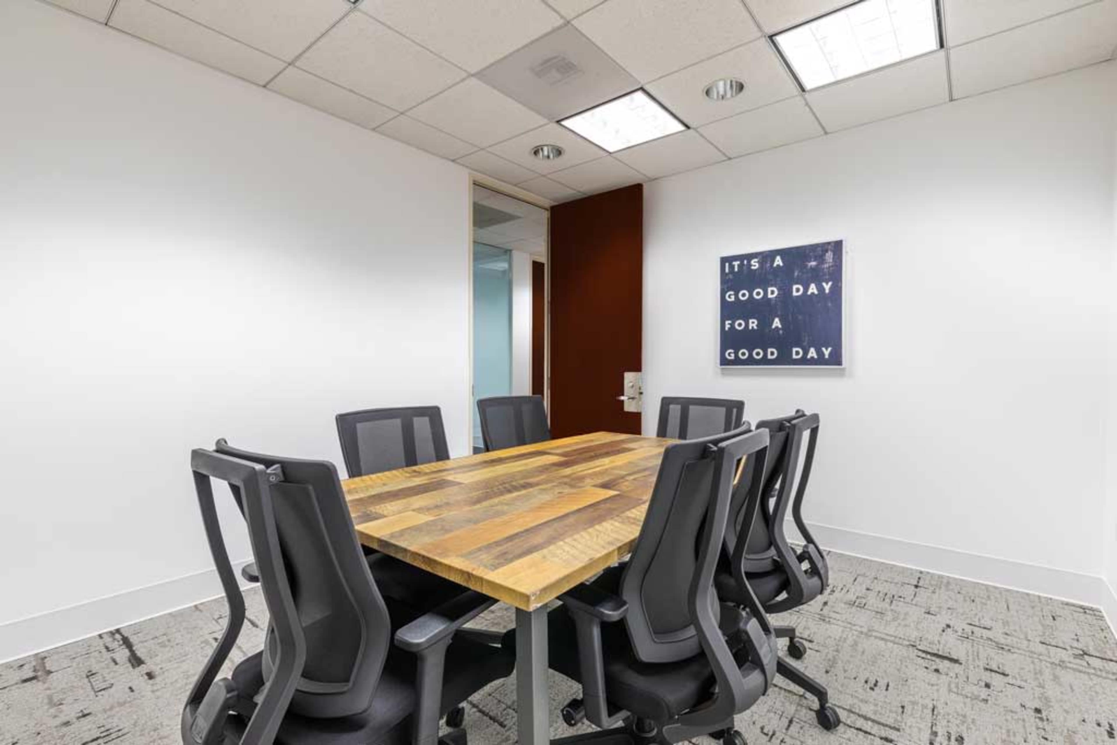 A meeting room features a wooden table surrounded by six black office chairs, with a motivational poster on the wall.