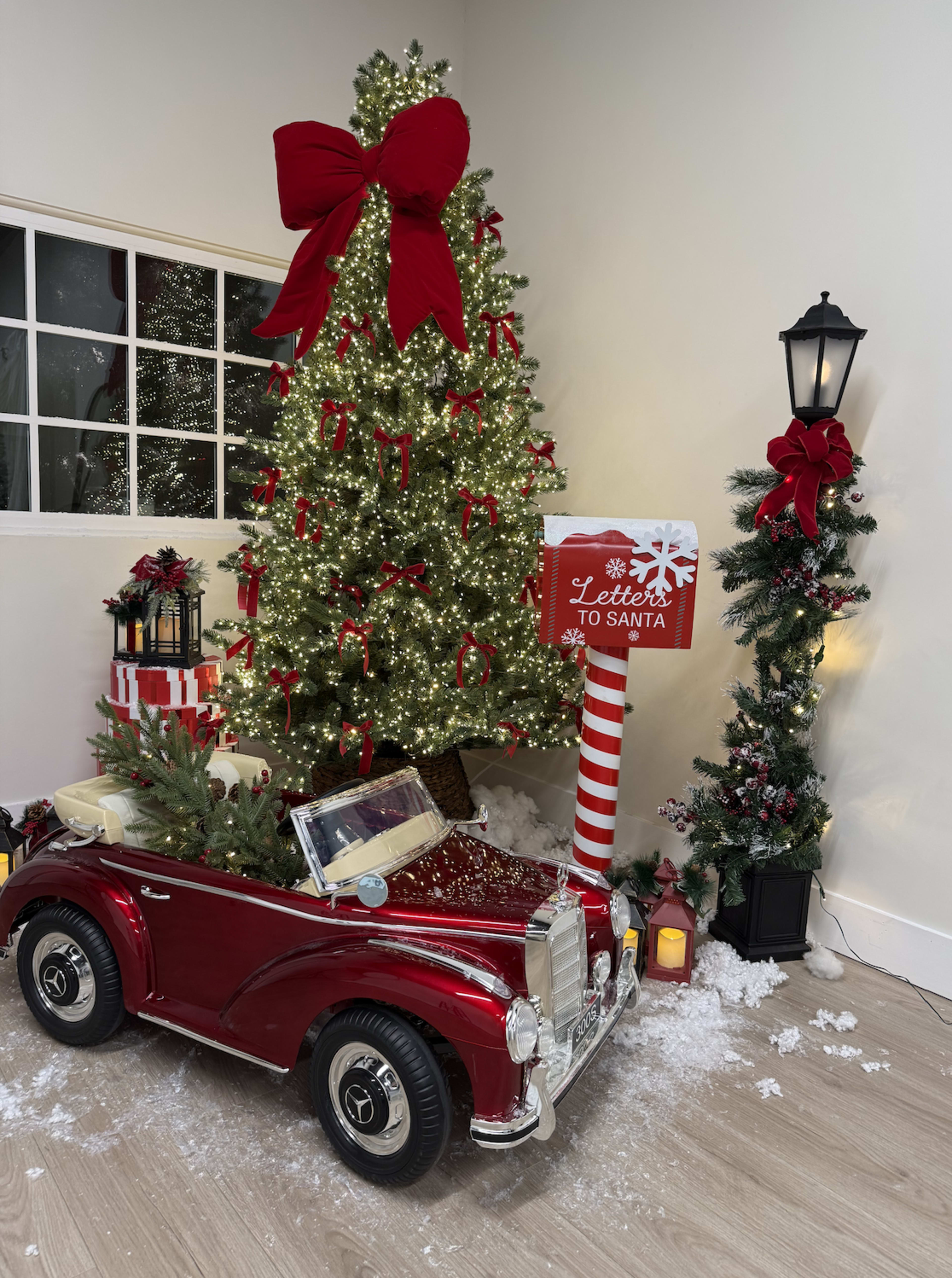 A festive scene features a small red toy car parked next to a Christmas tree adorned with lights and ribbons, alongside a mailbox labeled "Letters to Santa" and holiday decorations.
