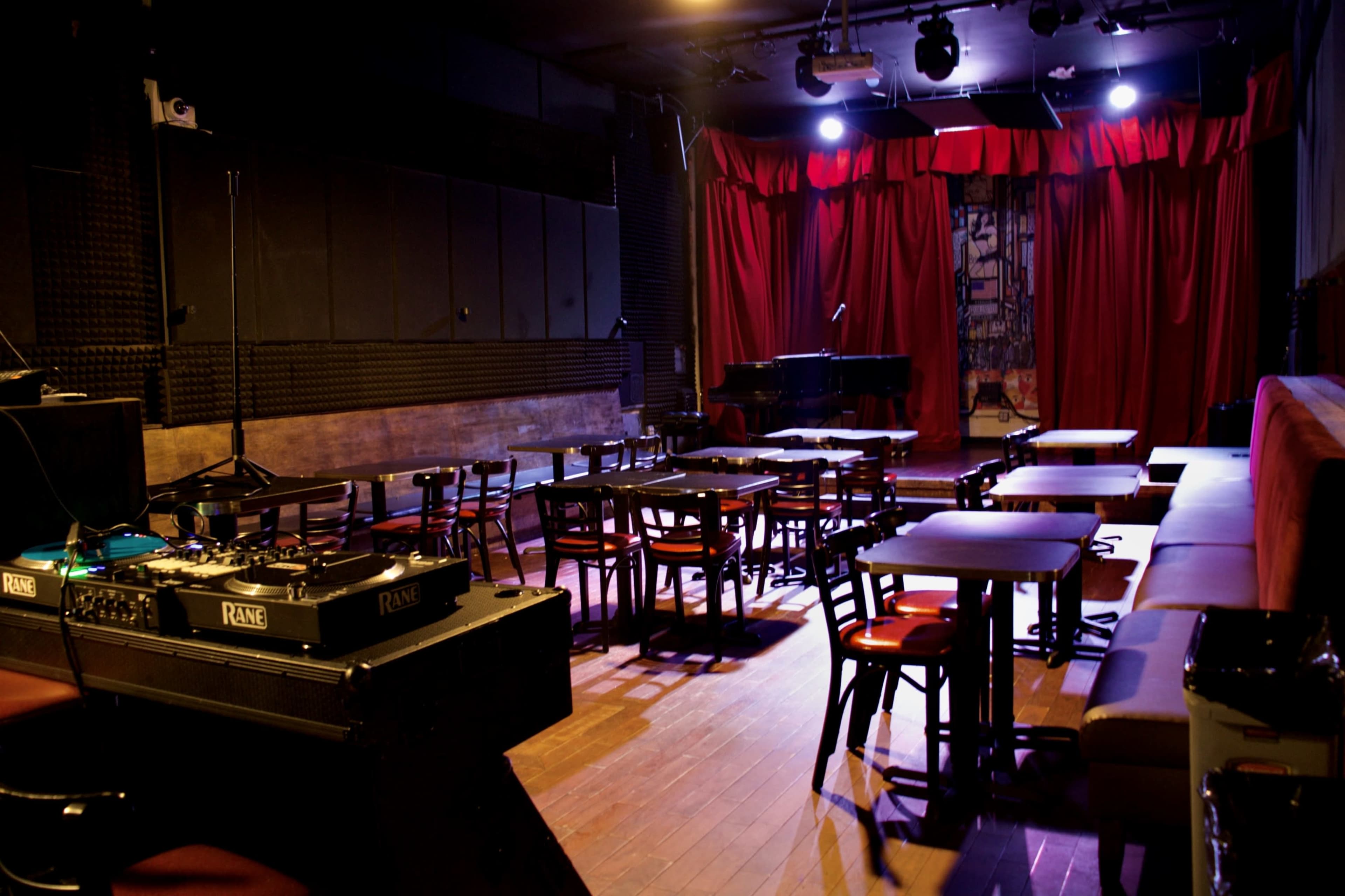 The image features a dimly lit music venue with red curtains, a small stage, and rows of tables and chairs arranged for an audience.