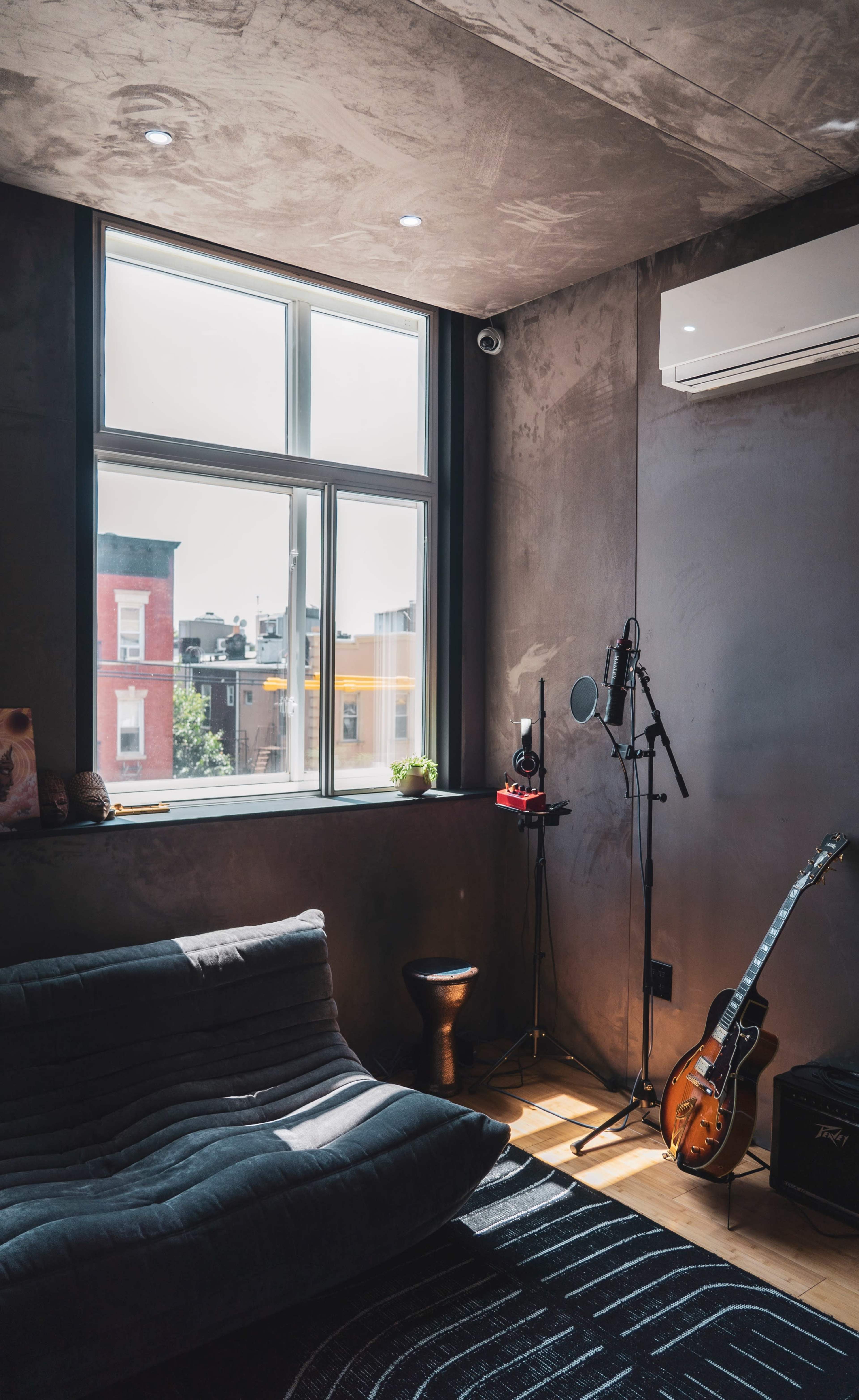 A sunlit room features a window, a guitar, a microphone setup, and a dark sofa against textured walls.