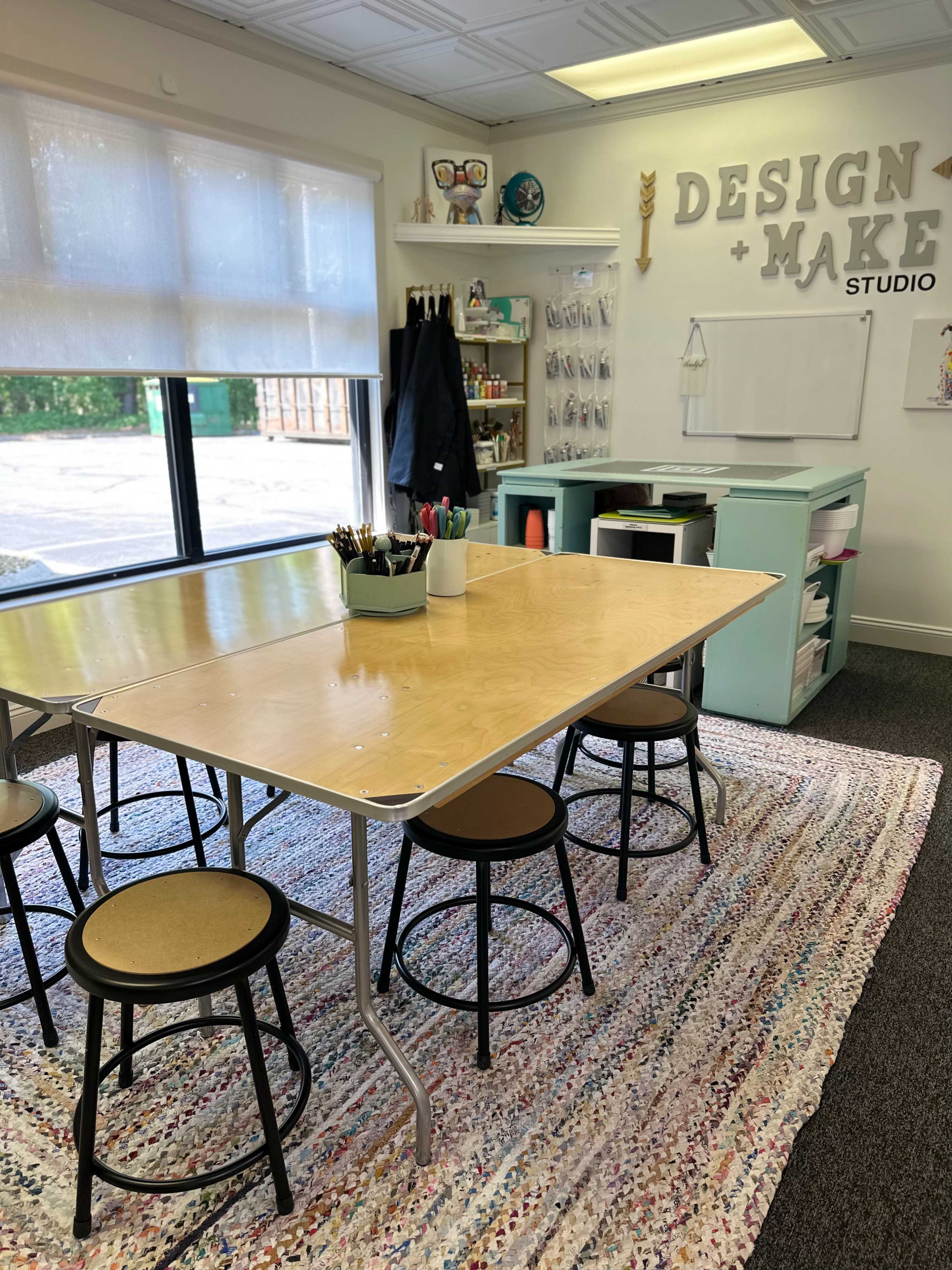 A bright design studio features a large wooden table surrounded by stools, with art supplies organized on shelves and a whiteboard mounted on the wall.