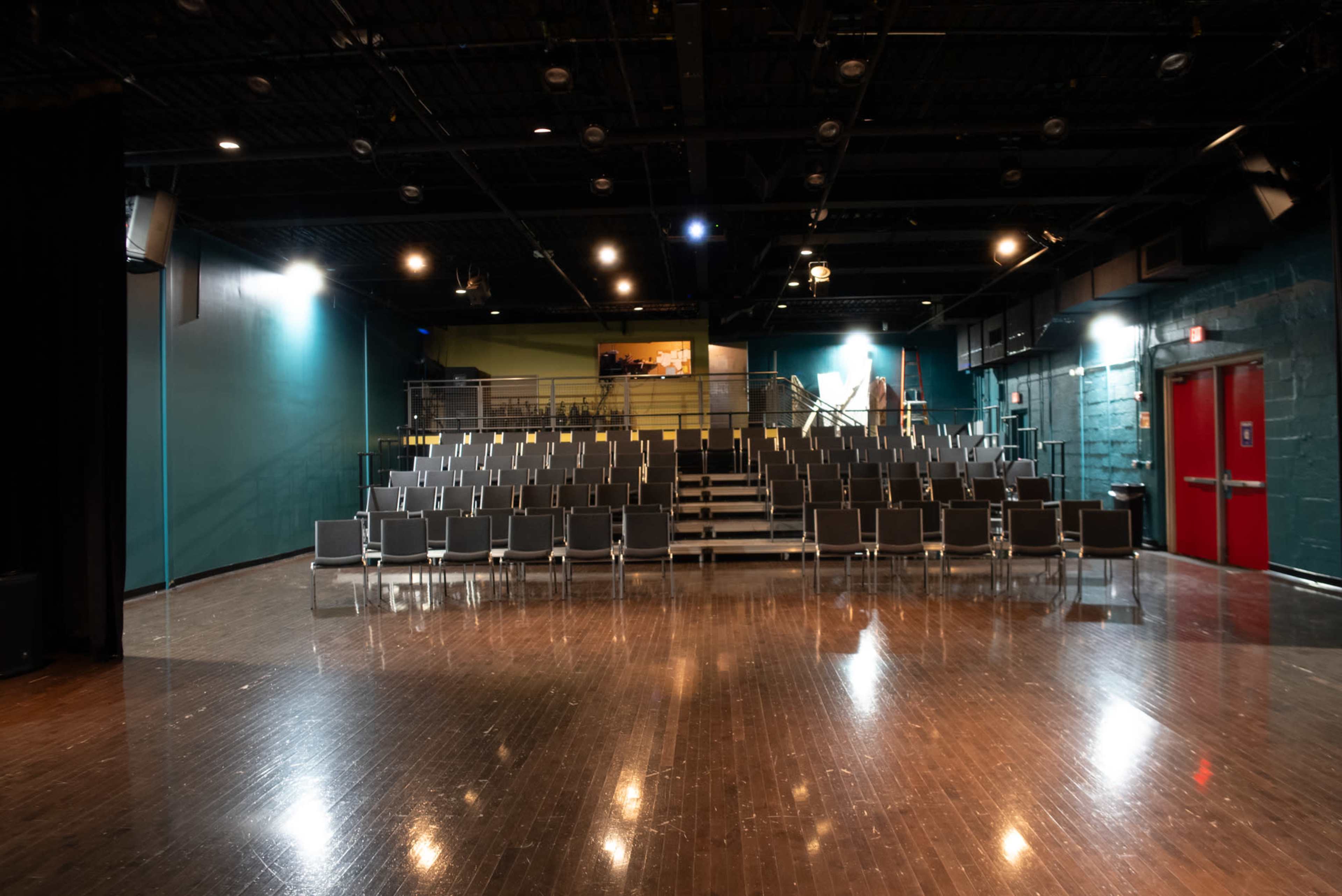 A spacious theater with a dark wooden floor, rows of empty chairs arranged in an audience area, and a red door in the background.