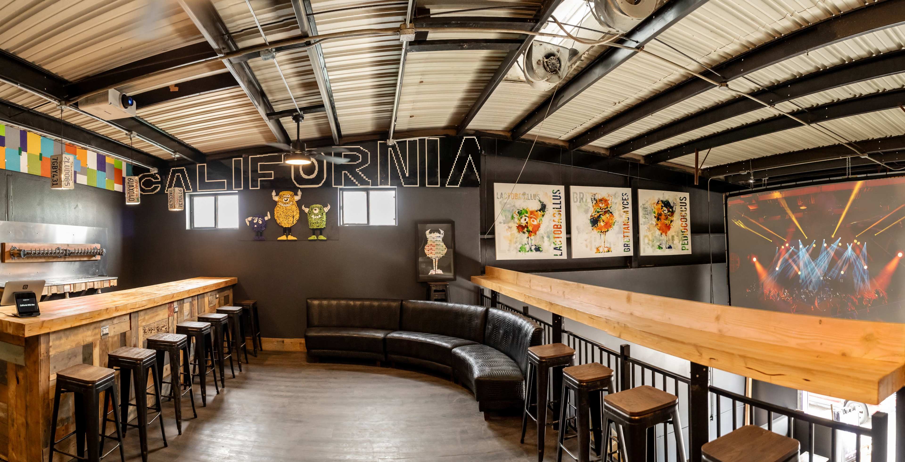 A modern bar area with a long wooden counter, black seating, and colorful wall art in a venue that features a "CALIFORNIA" sign and a large screen for displays.
