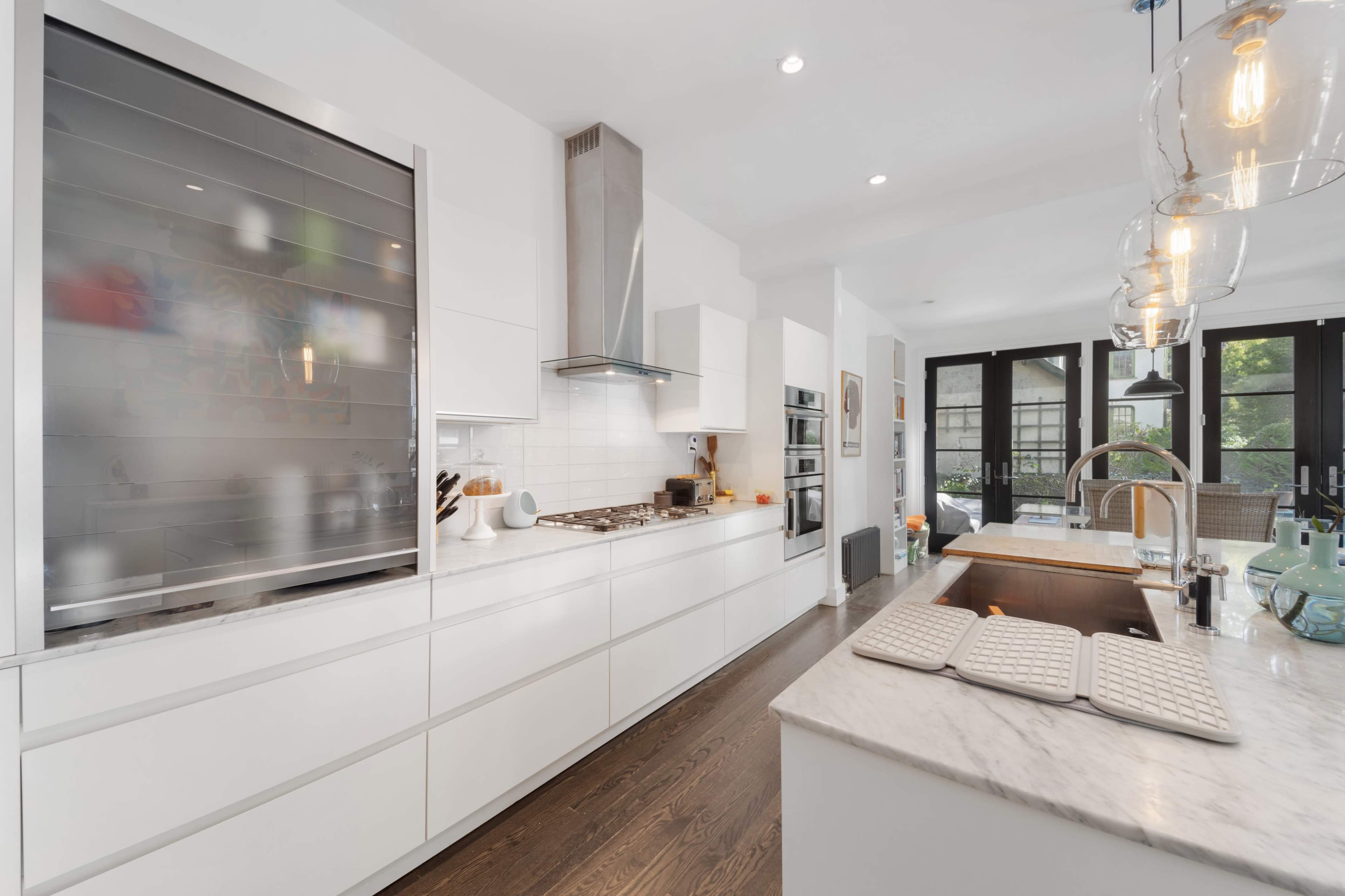 A modern kitchen features white cabinetry, a marble countertop, and a large stainless steel refrigerator, with a gas stove and pendant lighting.