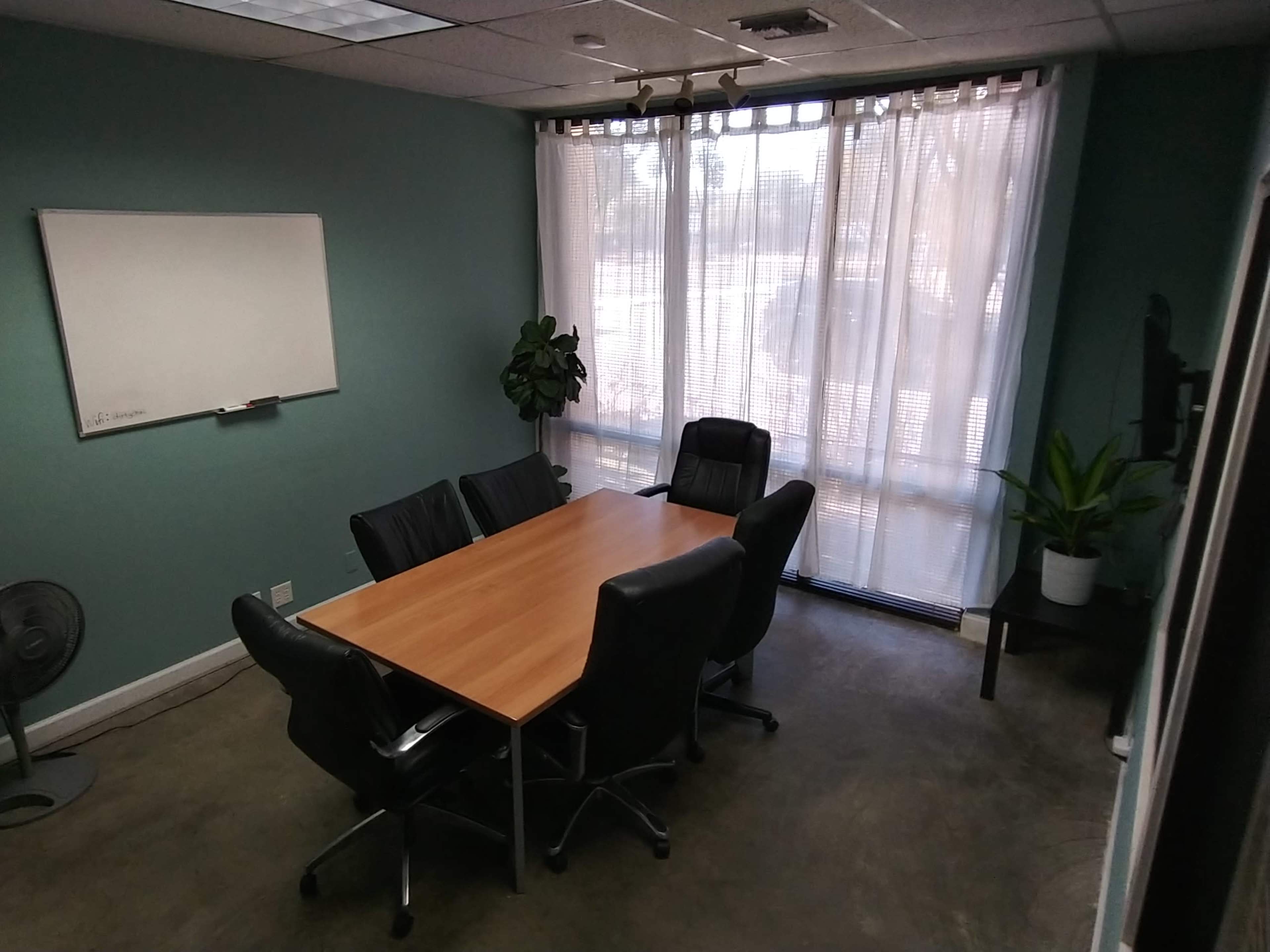 A conference room features a wooden table surrounded by six black leather chairs, with large windows covered by sheer curtains and a plant in the corner.