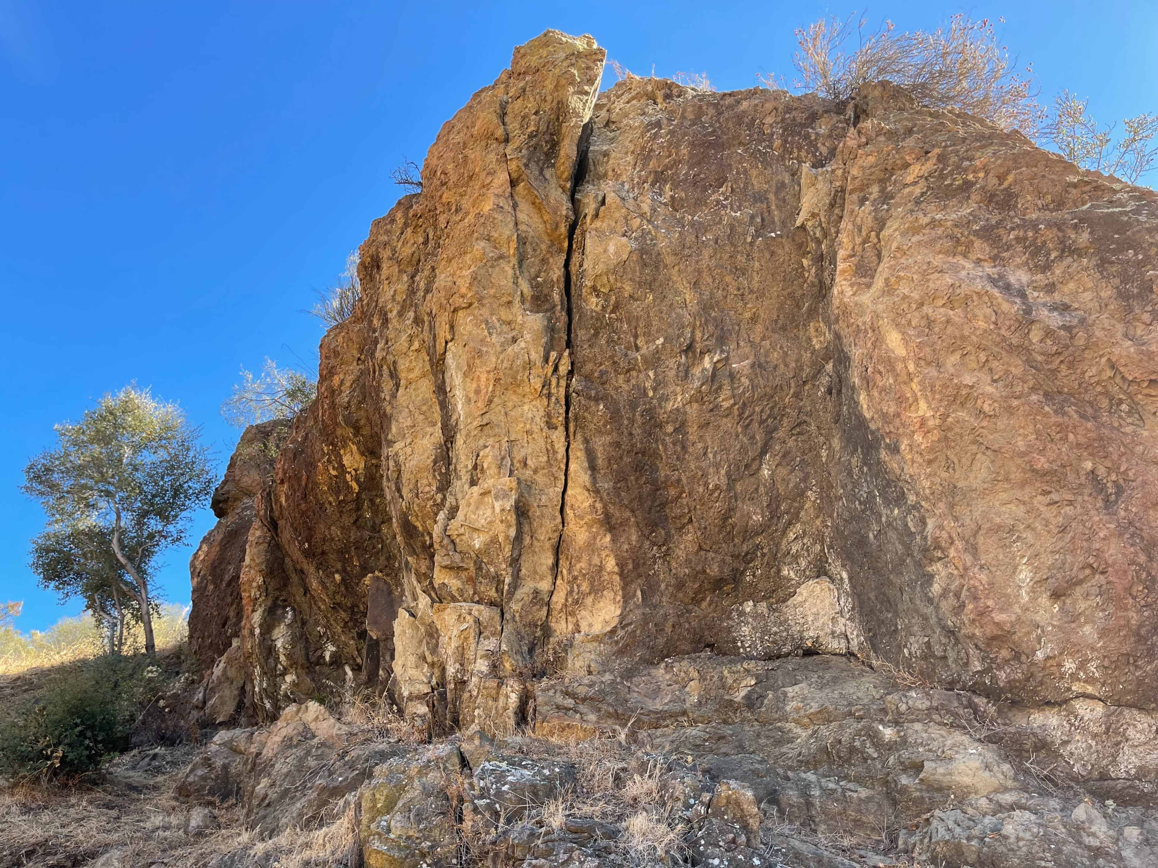 A large rock formation with a visible crack stands against a clear blue sky, surrounded by sparse vegetation.