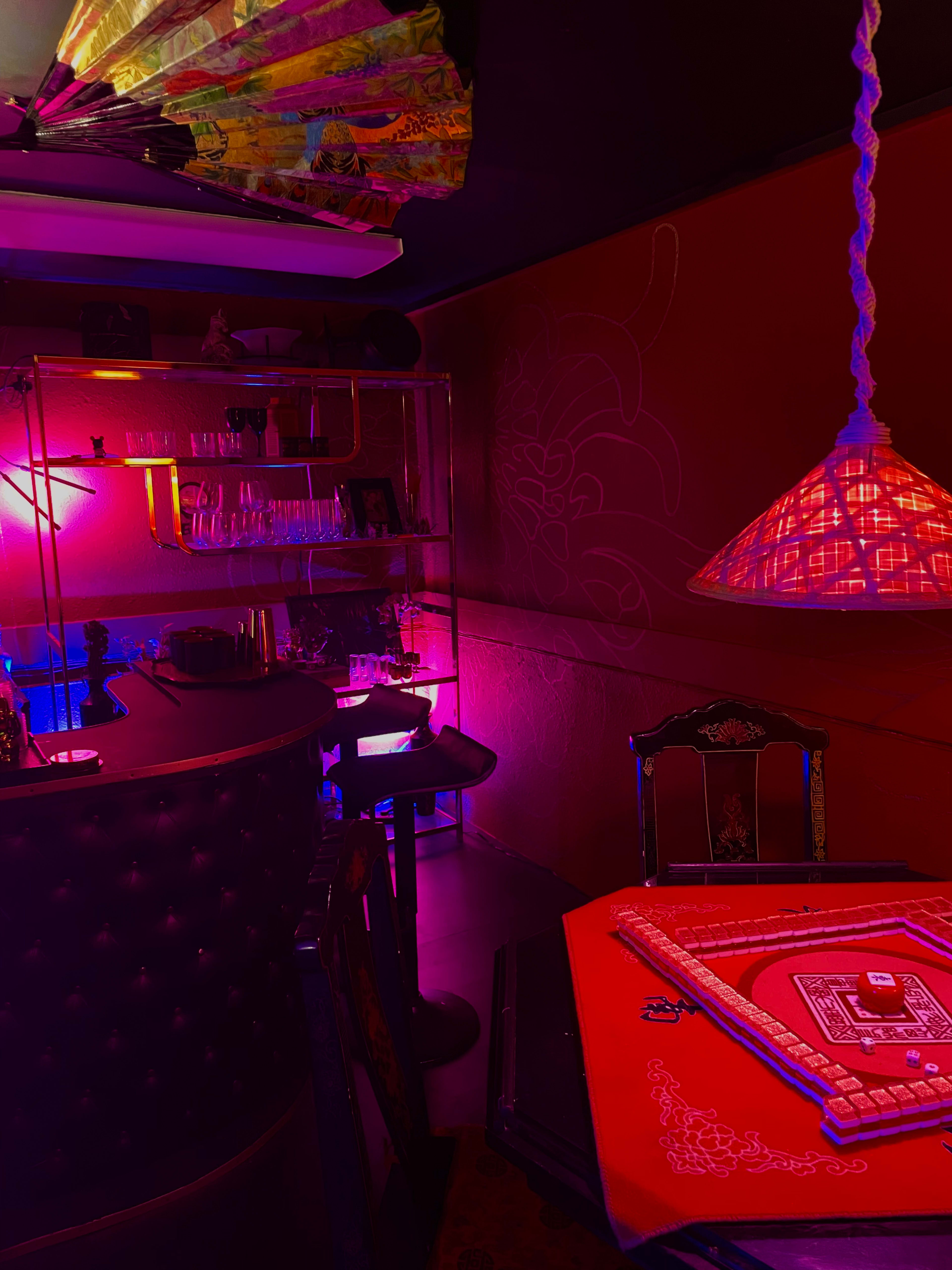 The image shows a dimly lit bar area with colorful lighting, a round table set for a game, and a decorative lamp overhead.