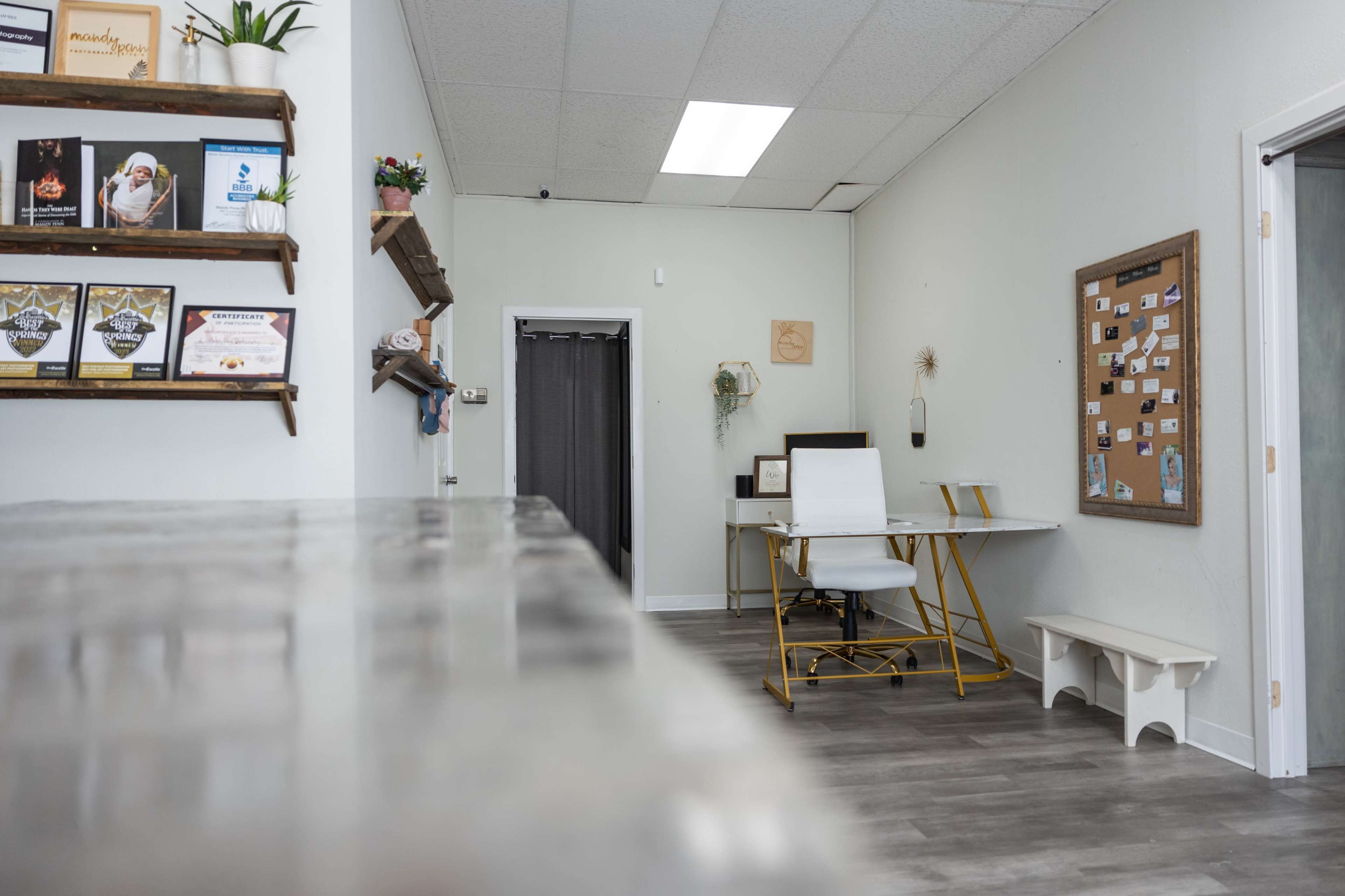 A bright, minimalist office space featuring a desk with a white chair, a bulletin board on the wall, and shelves displaying certificates and decorative items.