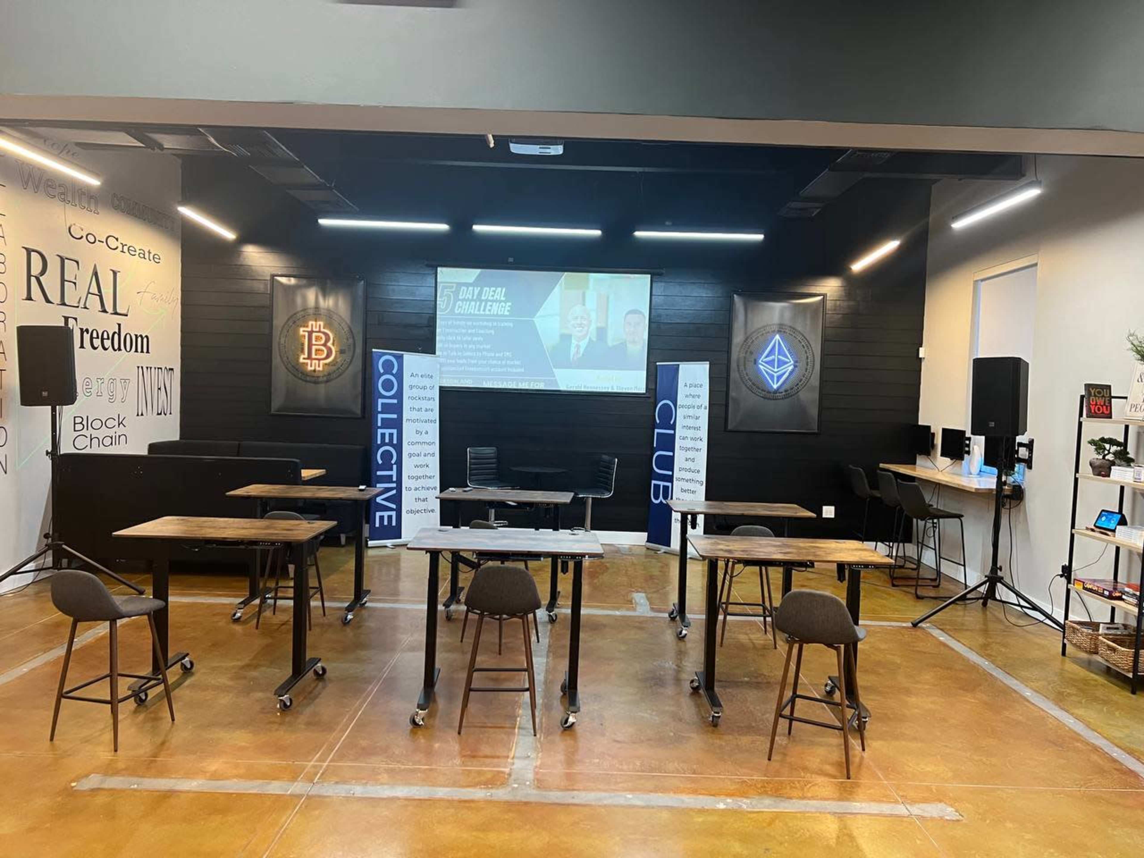 A modern indoor event space with several tables and chairs arranged, a large screen displaying a presentation, and branding related to blockchain and collective goals on the walls.
