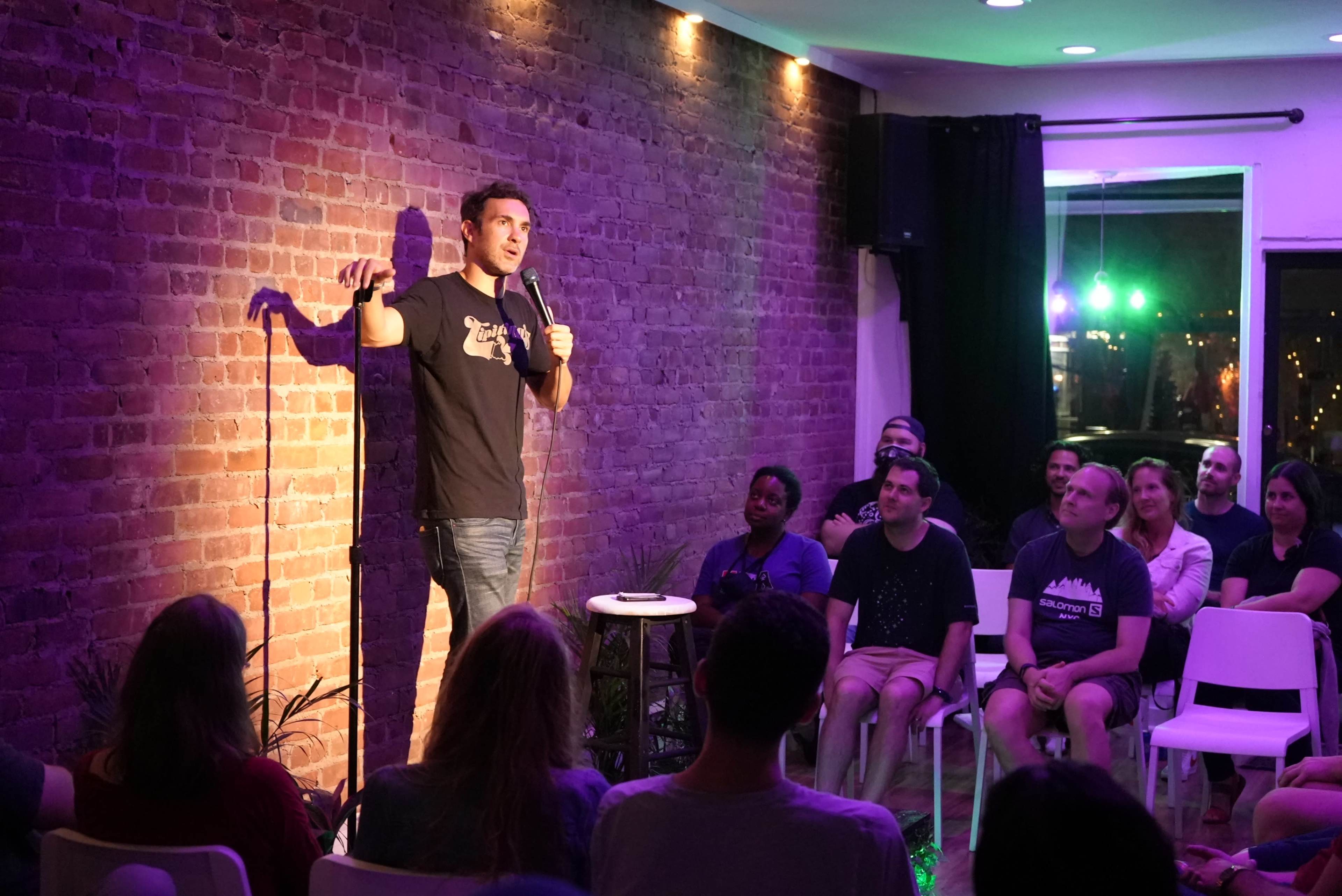 A comedian performs stand-up on stage in front of an audience seated in a dimly lit room with a brick wall backdrop.