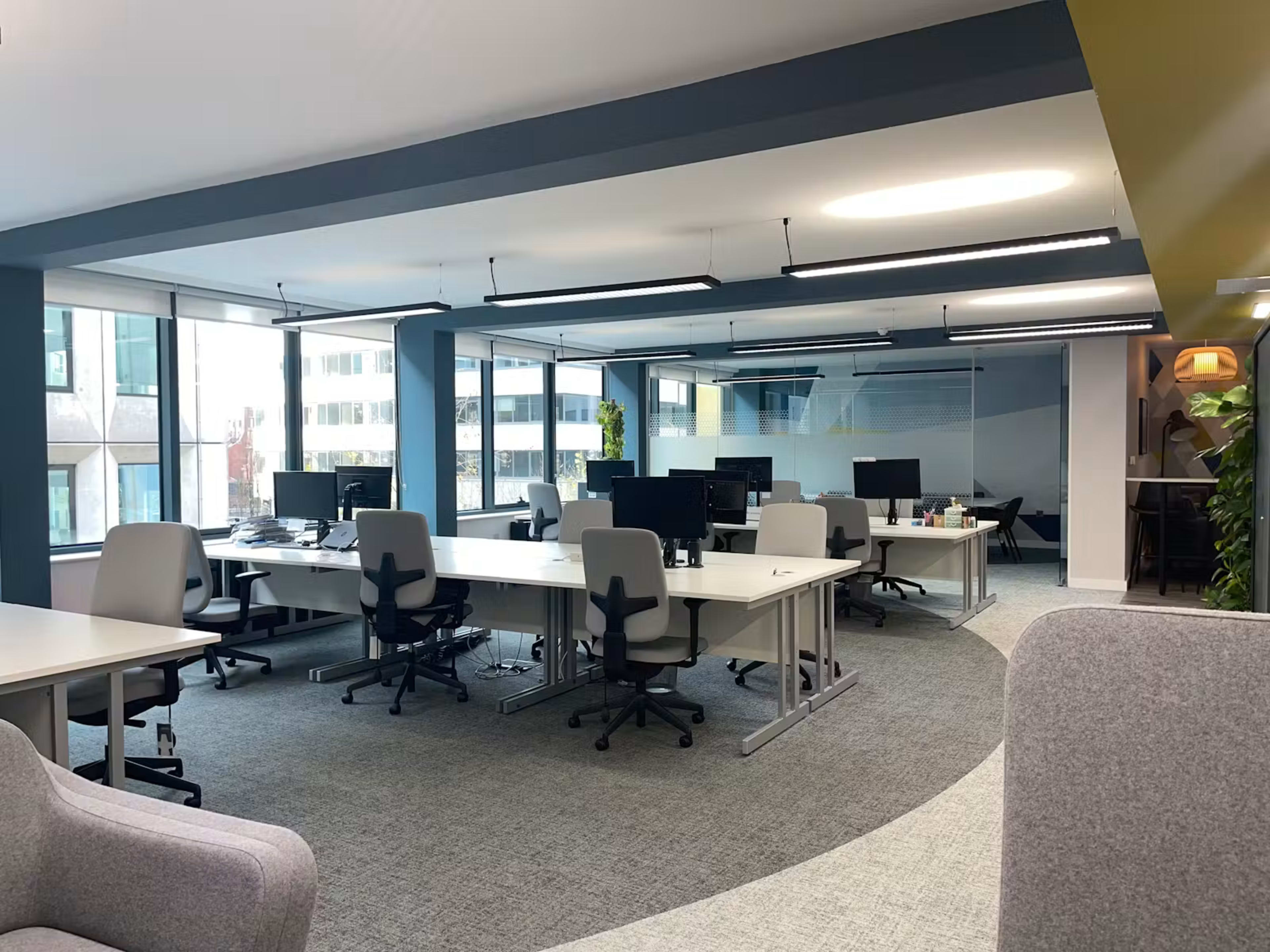 The image shows an open-plan office space with multiple desks equipped with computers and large windows allowing natural light to enter.