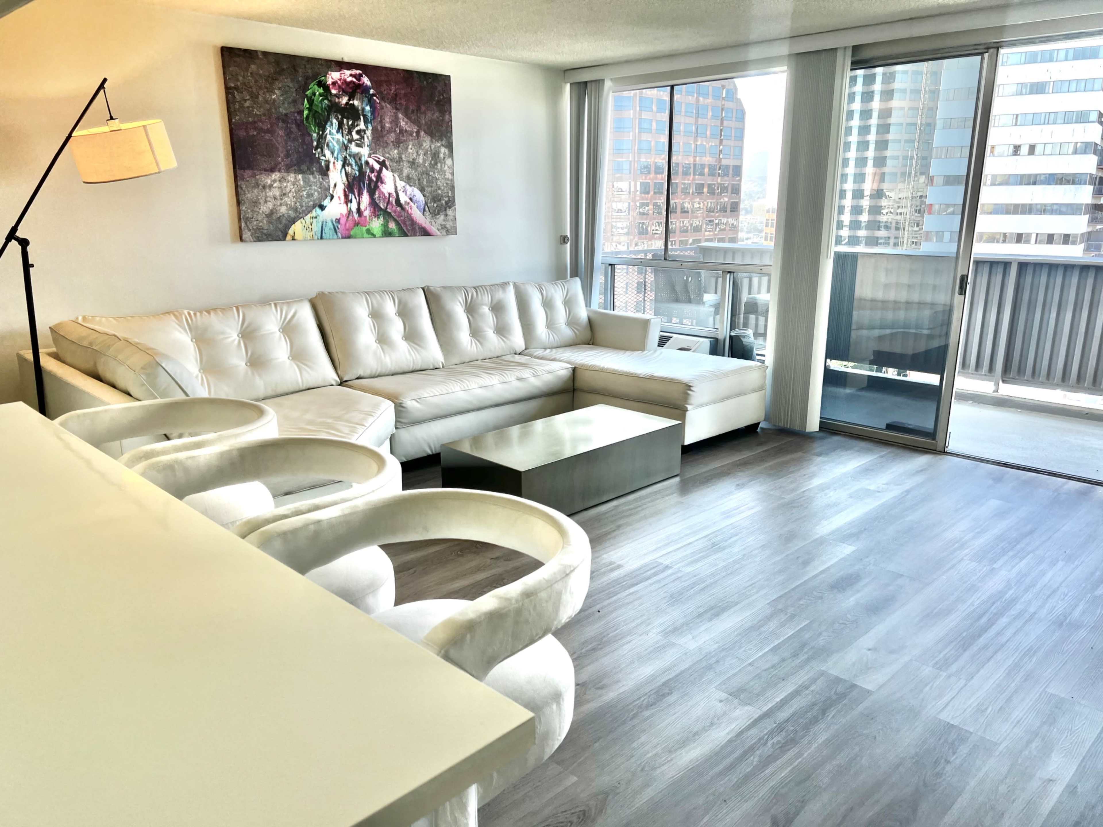 The image shows a modern, spacious living room with a white sectional sofa, a glass coffee table, and a large artwork on the wall, along with a view of city buildings through a floor-to-ceiling window.