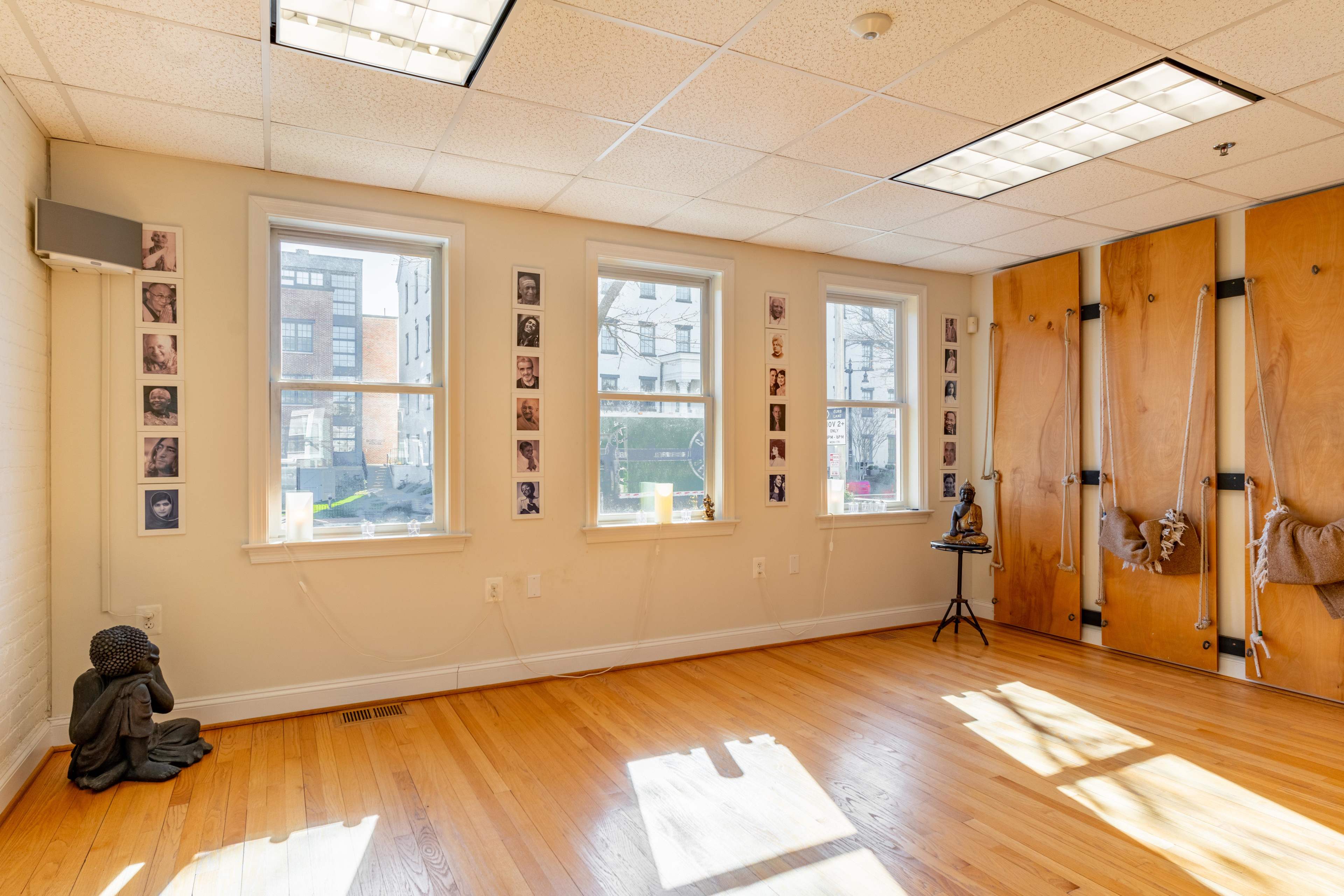 A spacious room with wooden flooring, featuring three windows, wall-mounted photographs, and a wooden partition on one side.