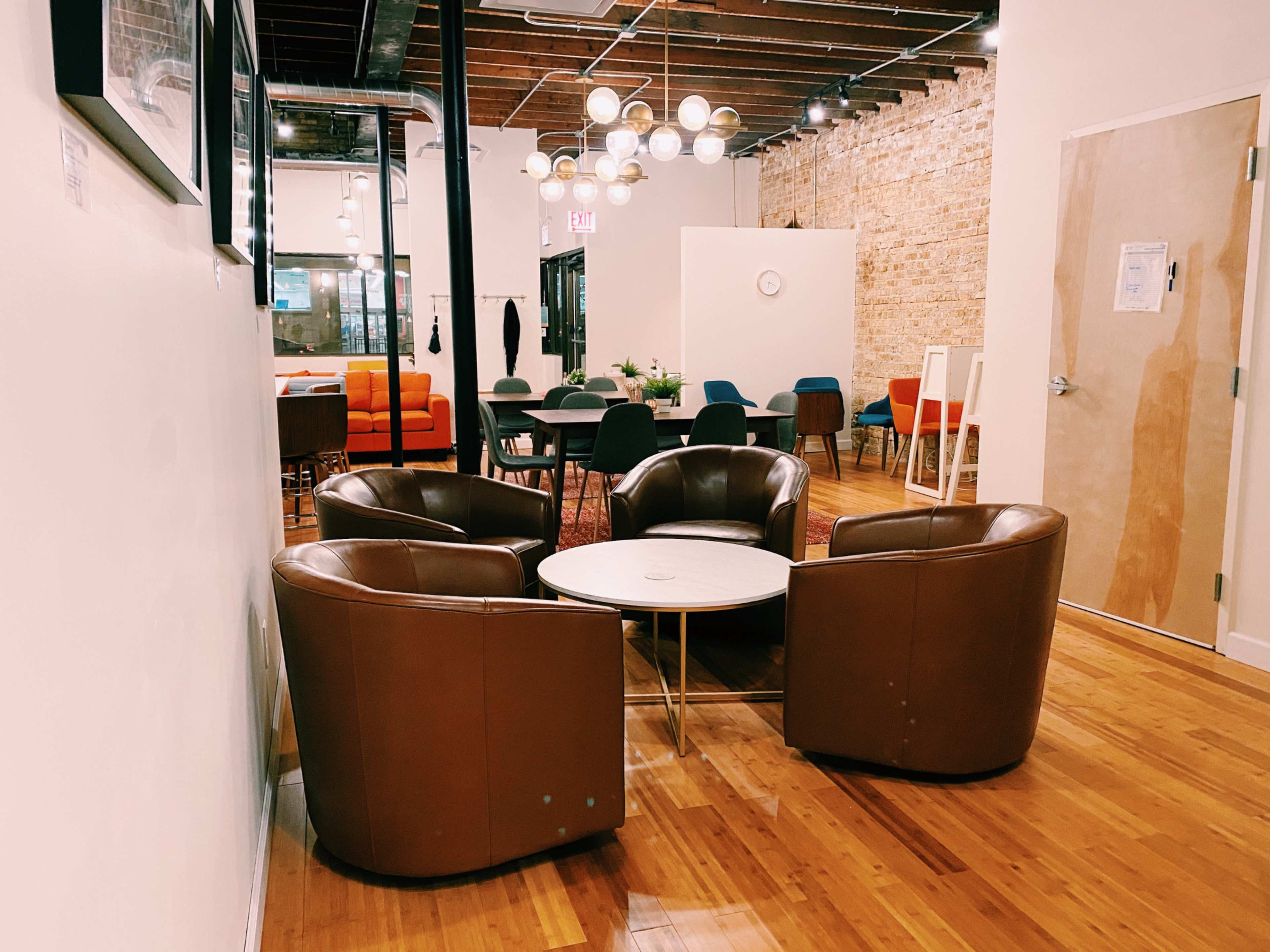 A modern lounge area featuring four brown chairs arranged around a circular coffee table, with a mix of dining tables and chairs in the background.