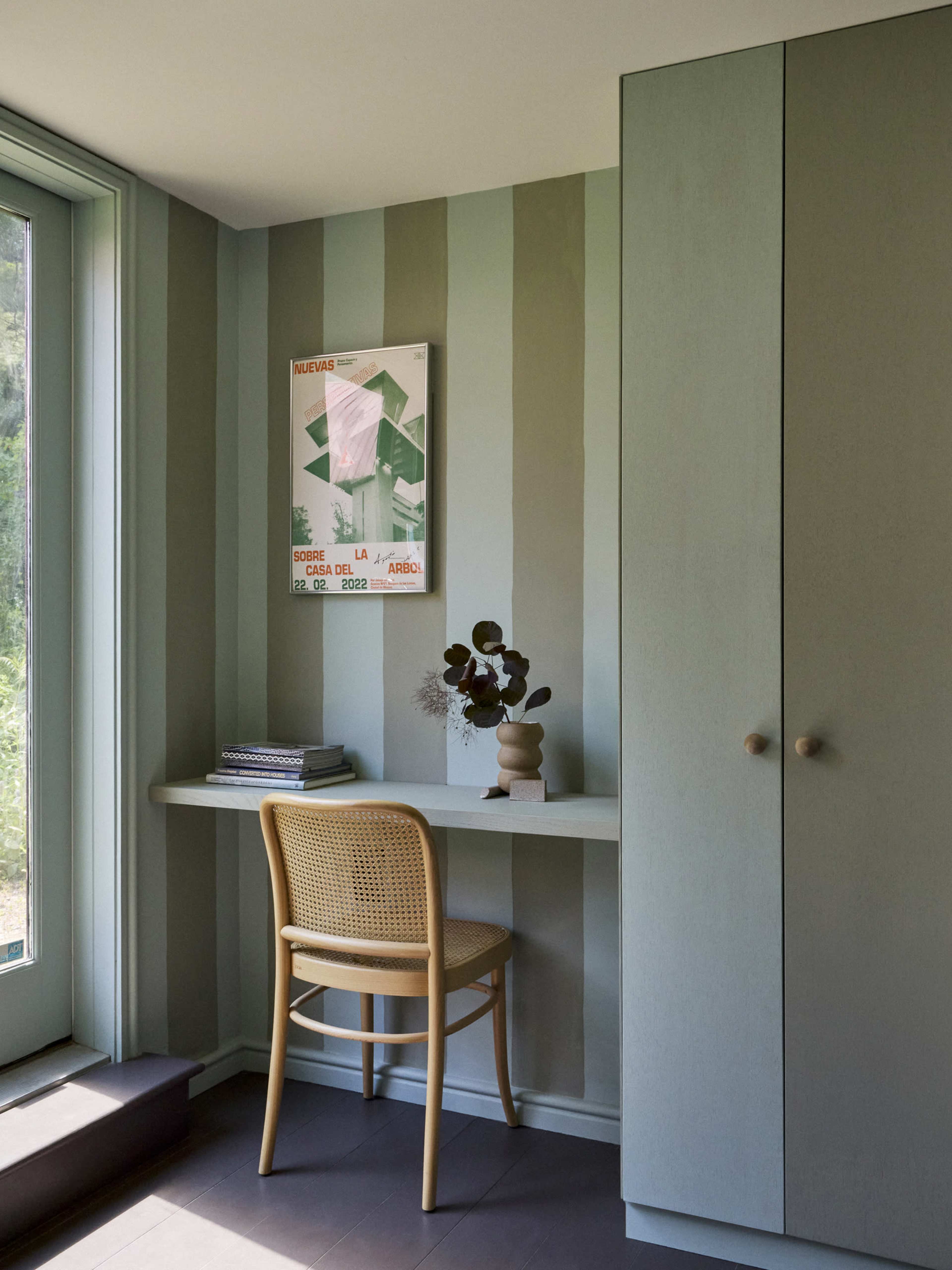 The image shows a corner of a room featuring striped green walls, a small desk with a chair, and a closed cabinet.