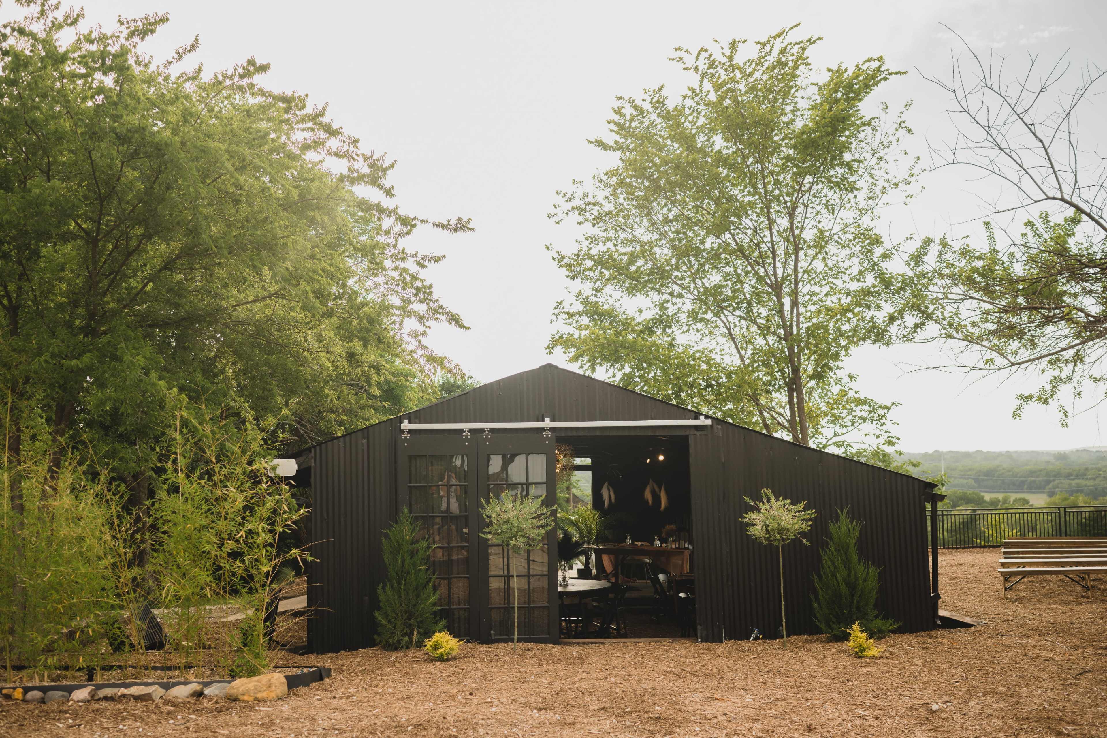 A modern black structure with large glass doors is surrounded by greenery and gravel pathways.