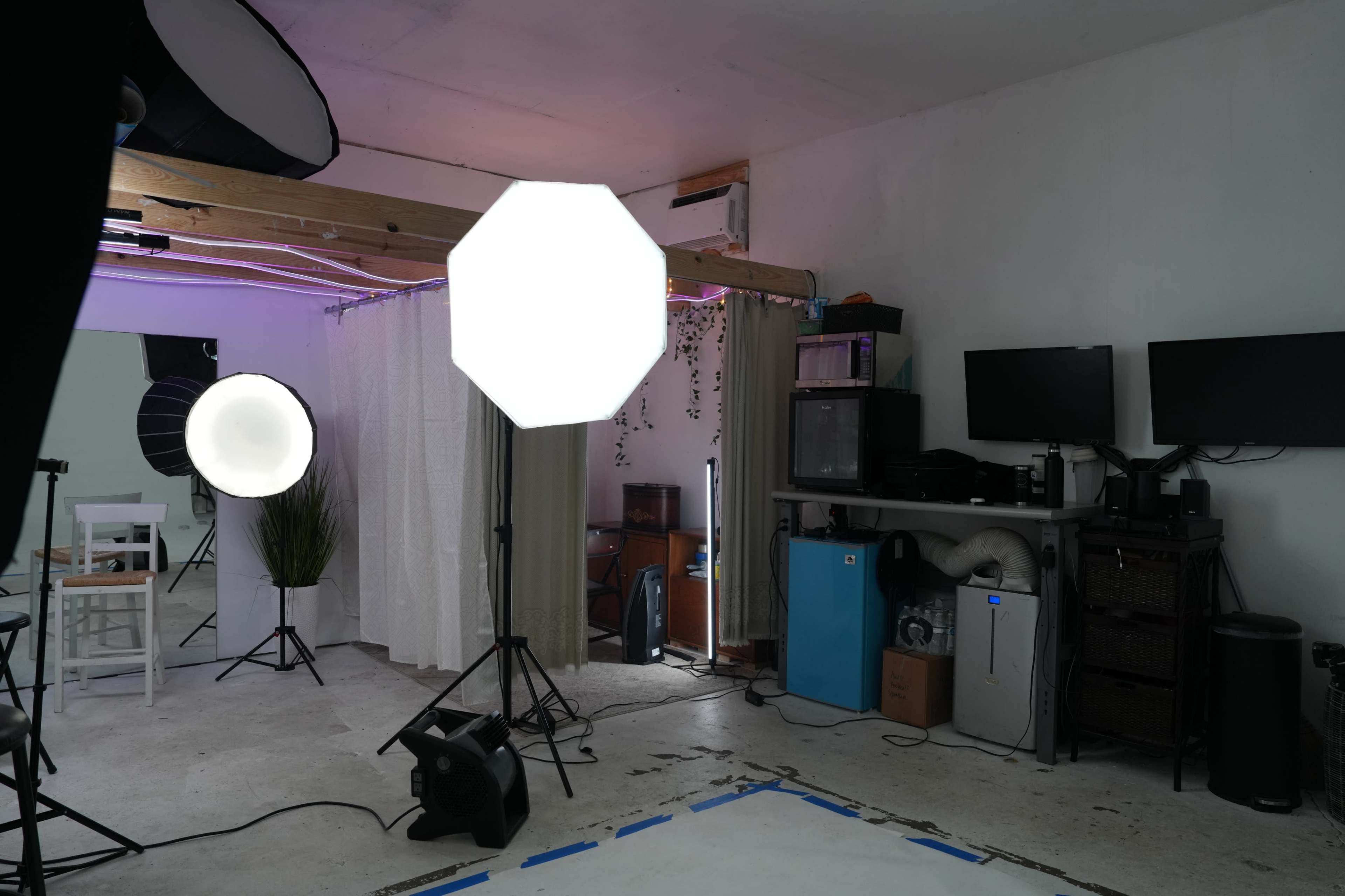 A photography studio is set up with various lighting equipment and a workstation featuring computers and appliances against a white wall.