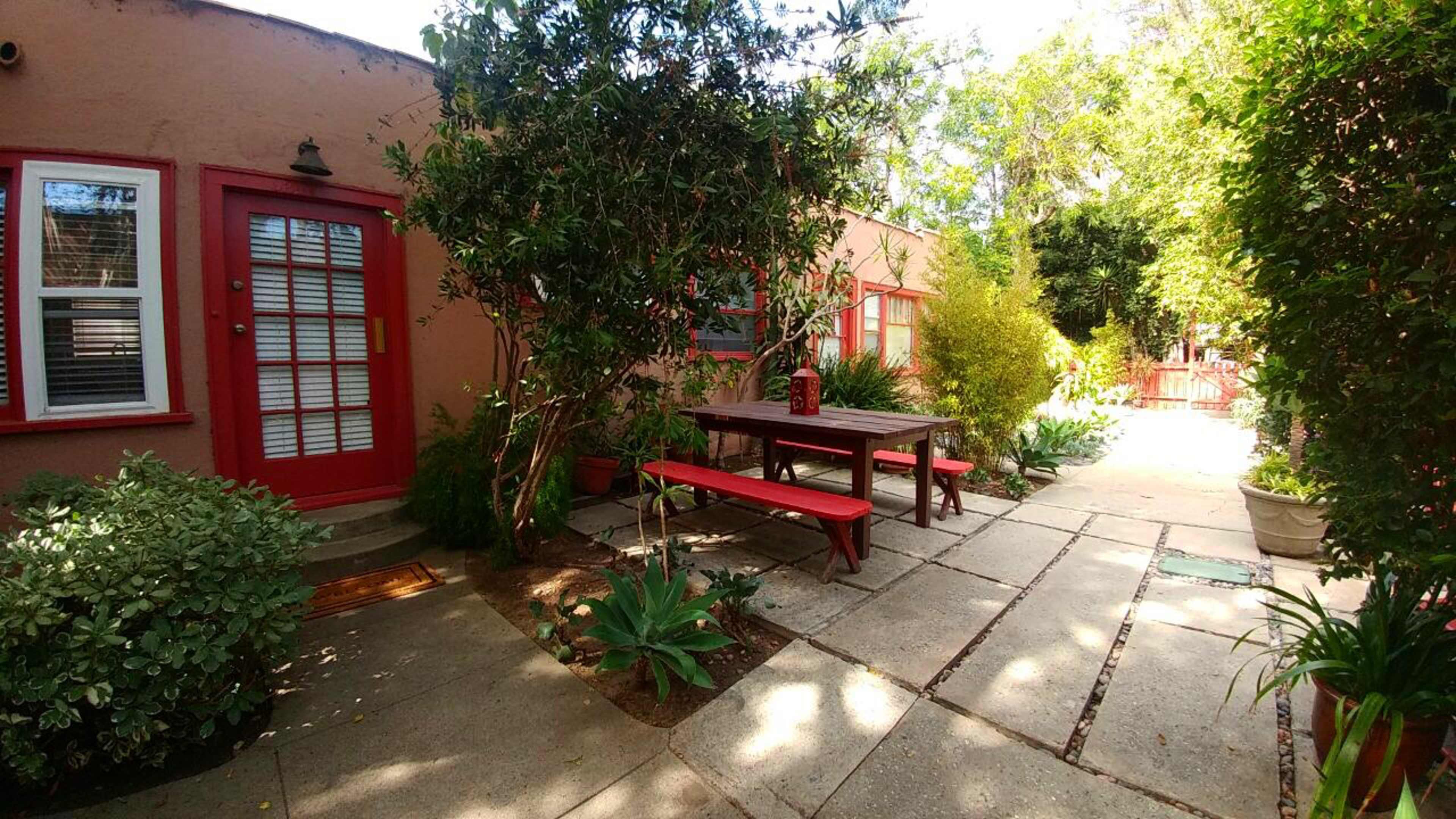 A patio area with a red picnic table surrounded by greenery and potted plants, leading to a building with a red door.