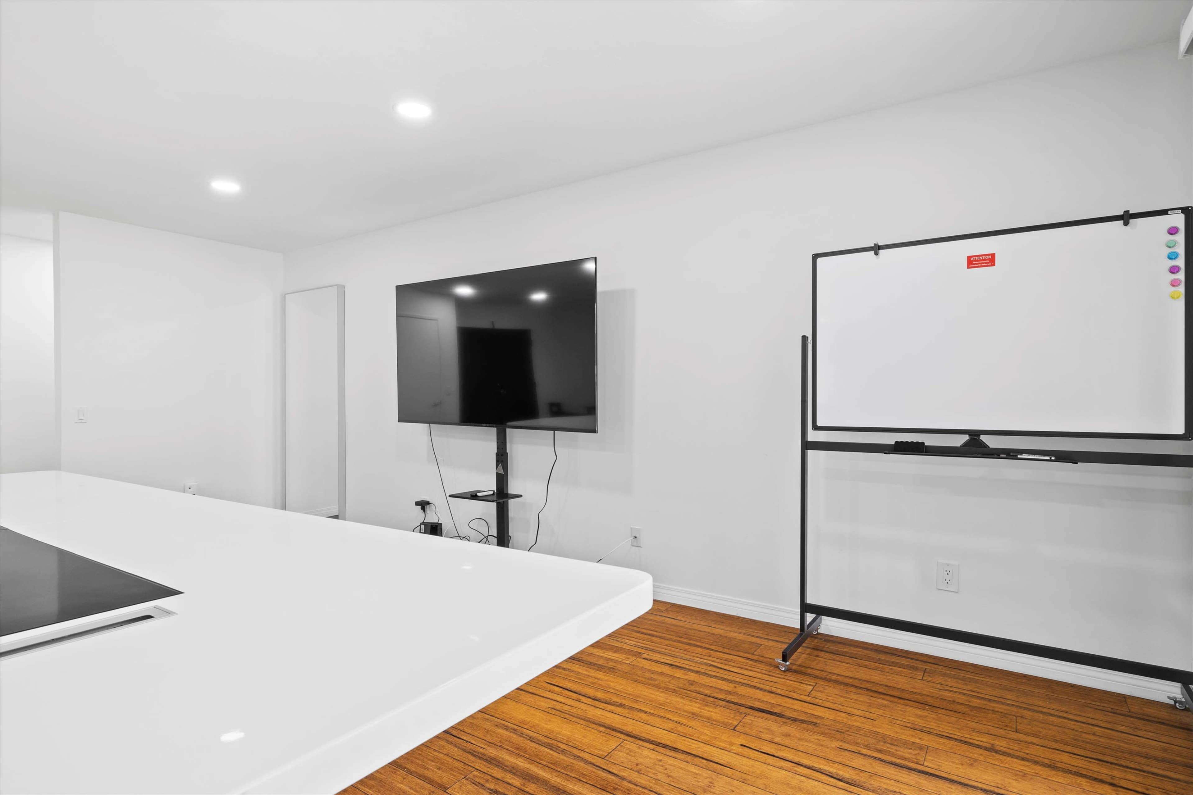 The image shows a modern, minimalist conference room with a large flat-screen TV mounted on the wall and a whiteboard on a movable stand next to a sleek, white table.