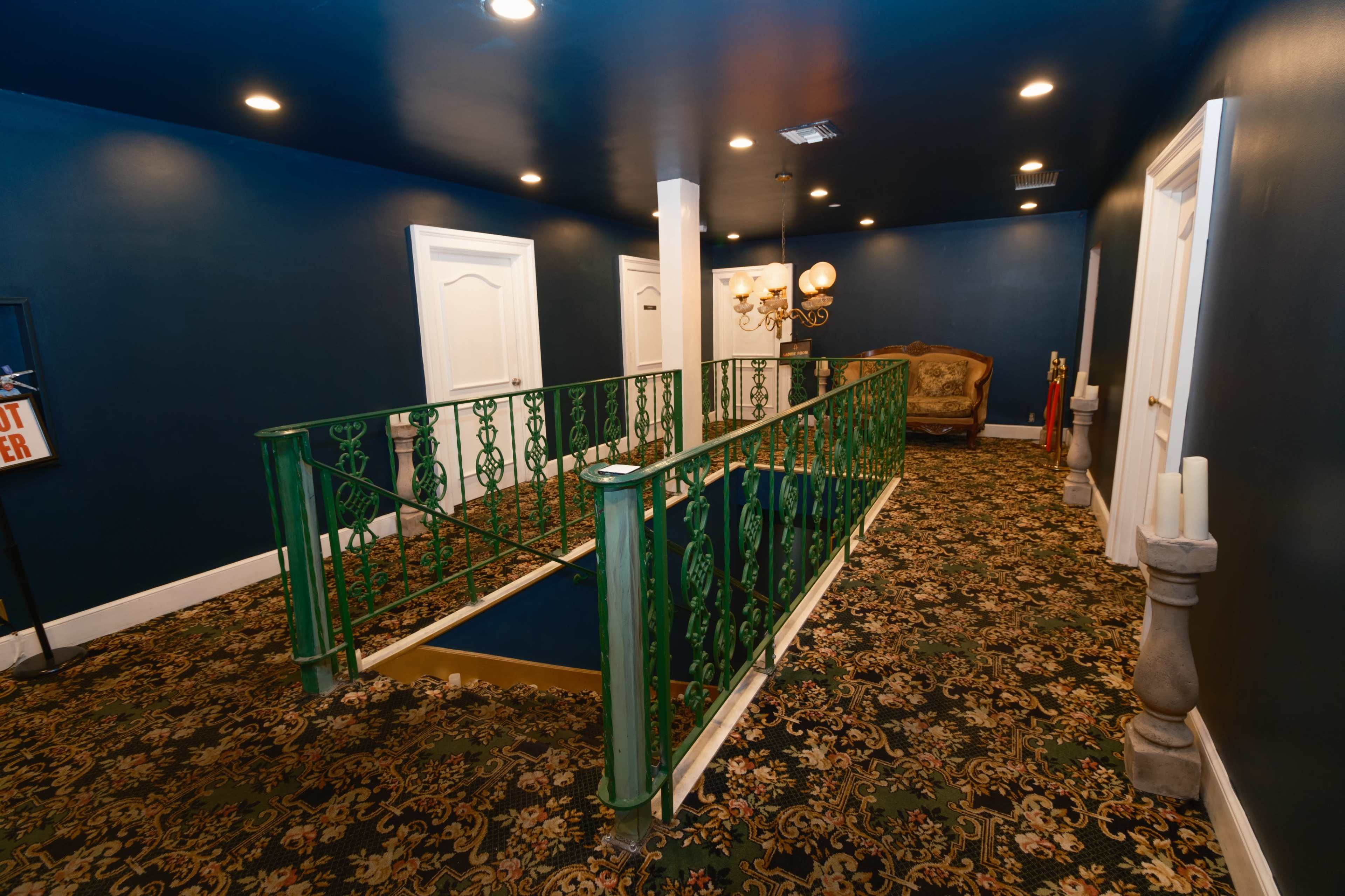 The image shows a hallway with dark blue walls and a green railing, leading to a seating area with a decorative chair and various light fixtures.