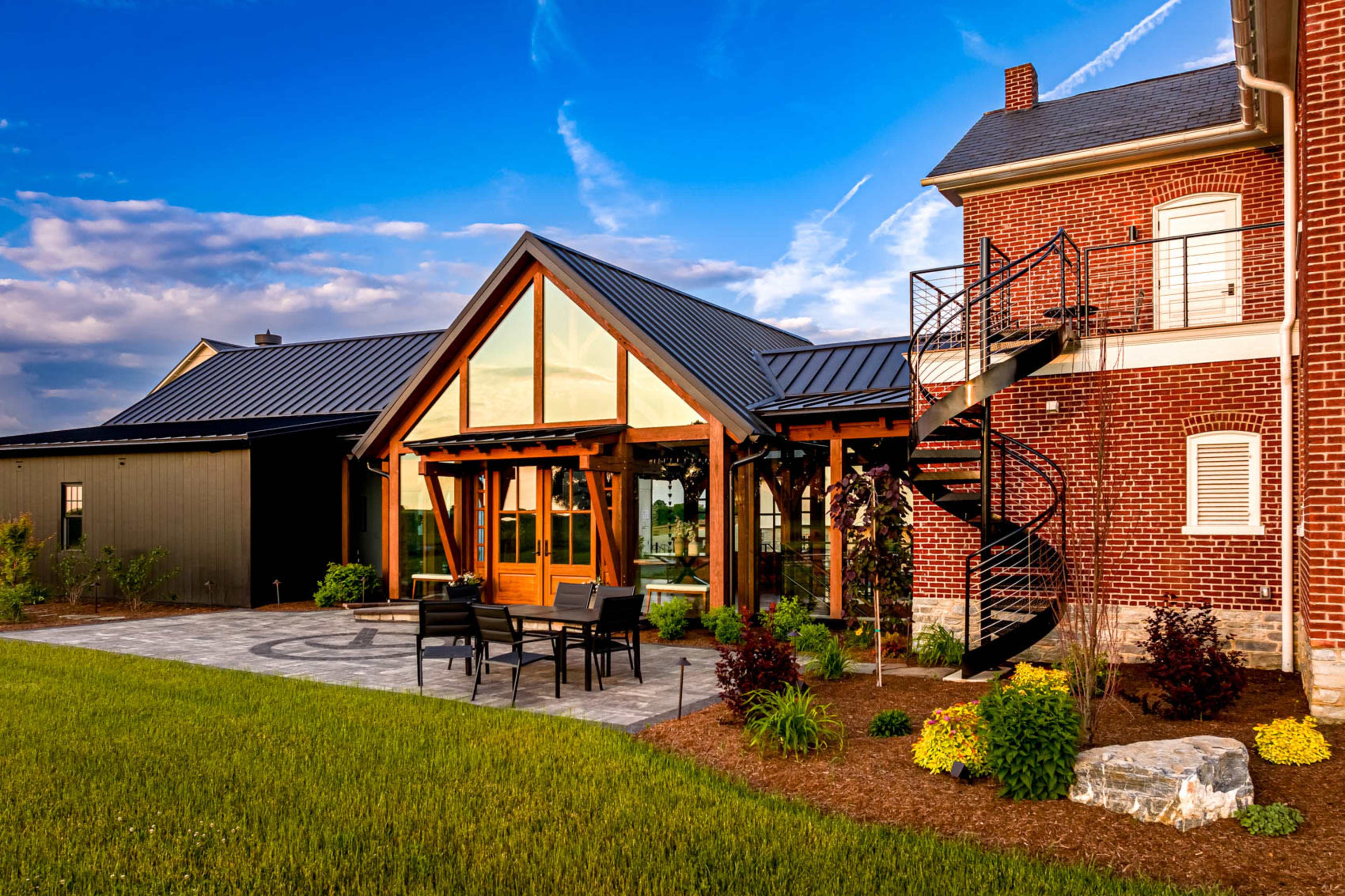 A modern brick house features large glass windows, a spiral staircase, and a patio with black outdoor furniture surrounded by greenery.