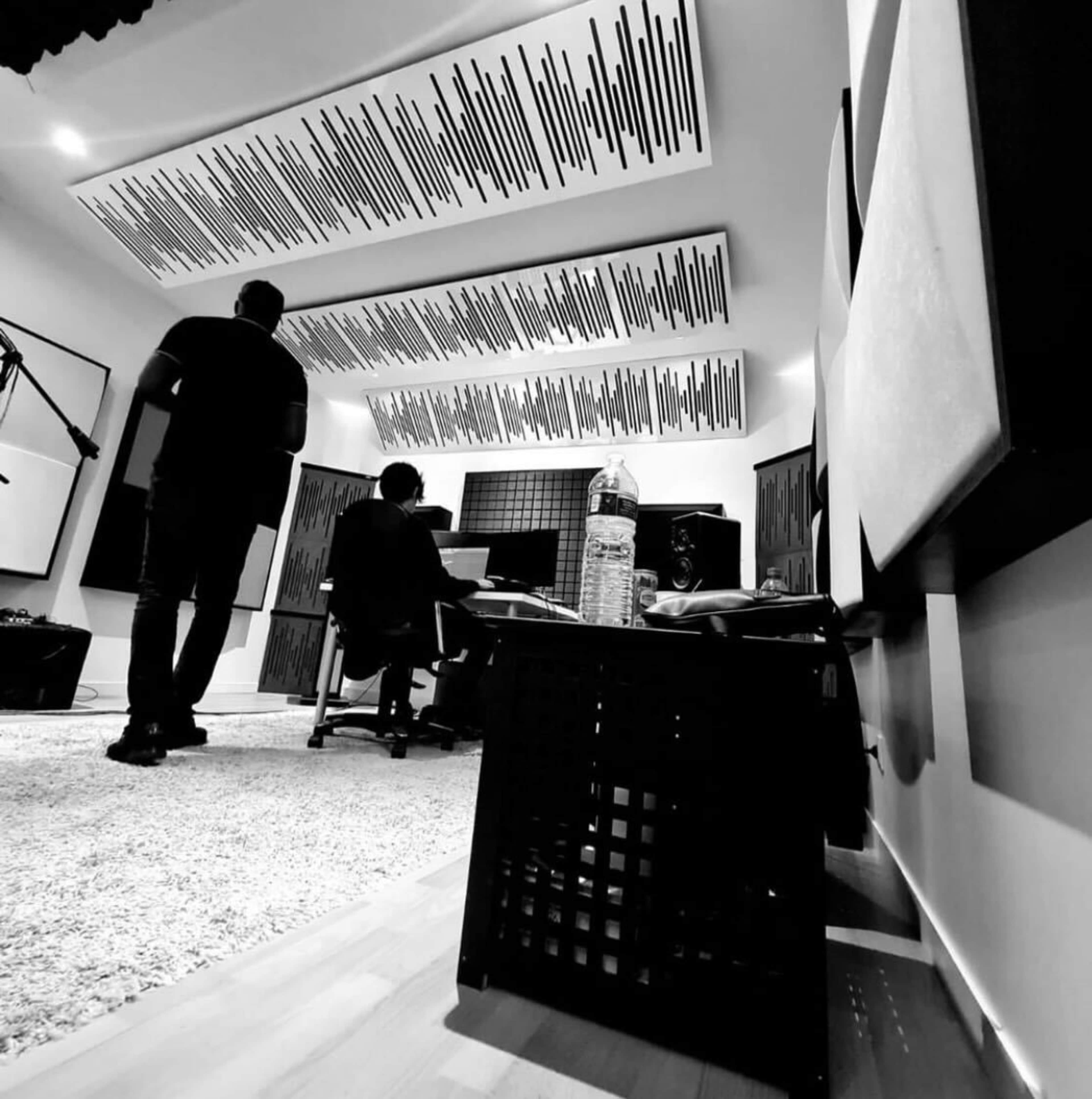 A black and white interior of a recording studio with two people, one standing and one sitting at a desk, surrounded by soundproofing panels and audio equipment.