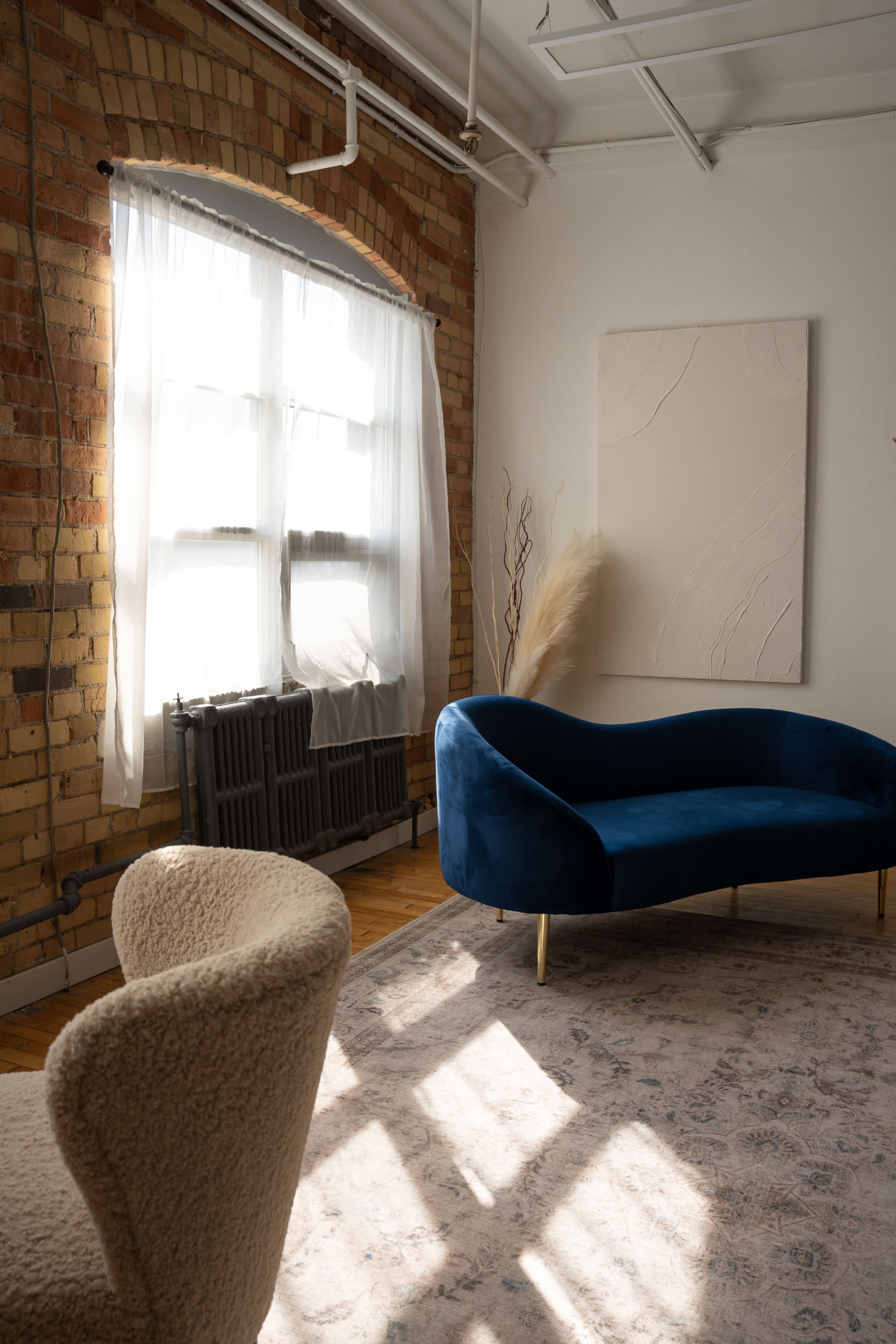 The image shows a bright, minimalist room with a blue couch and a light-colored armchair, featuring large windows that let in natural light.