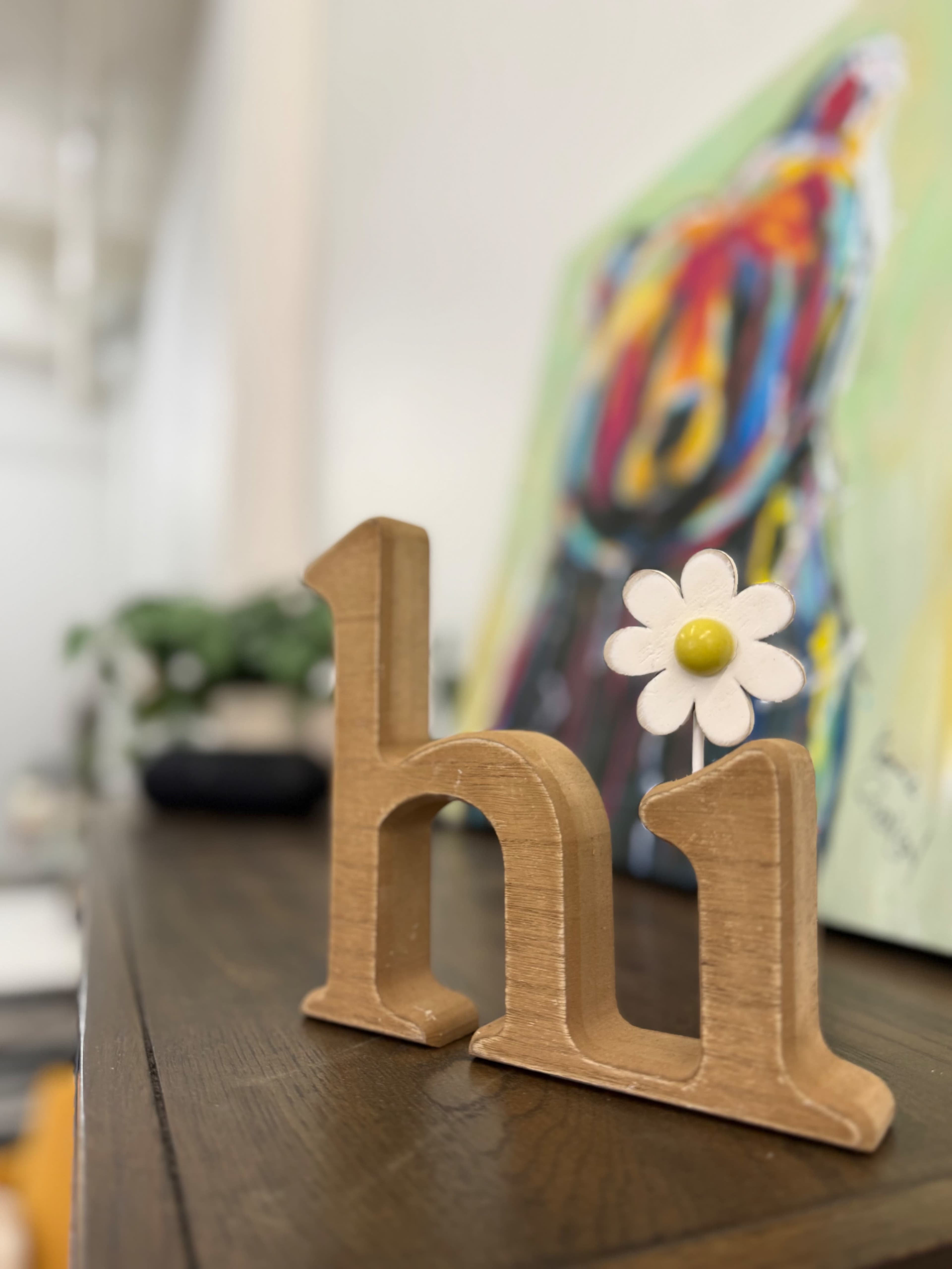 A wooden decoration displaying the word "hi" with a small flower attached is positioned on a shelf beside a colorful painting.