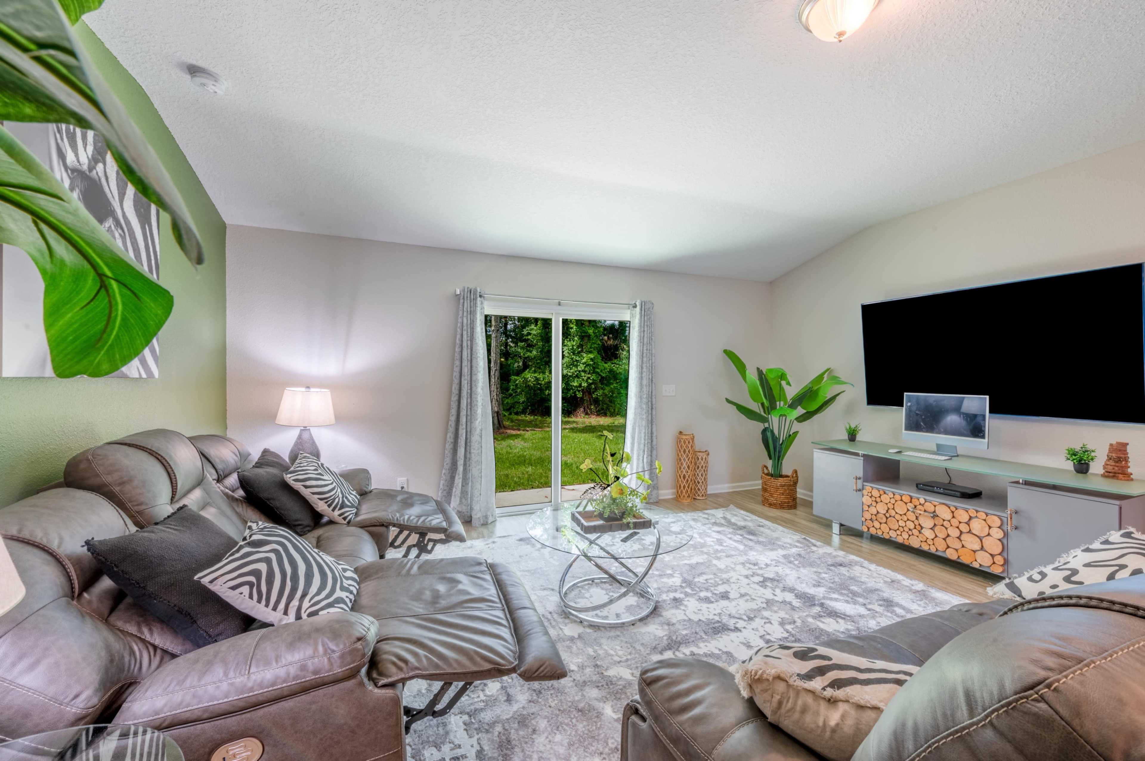 A living room features dark leather recliners, a glass coffee table, a large TV mounted on the wall, and a sliding door that opens to a green outdoor view.