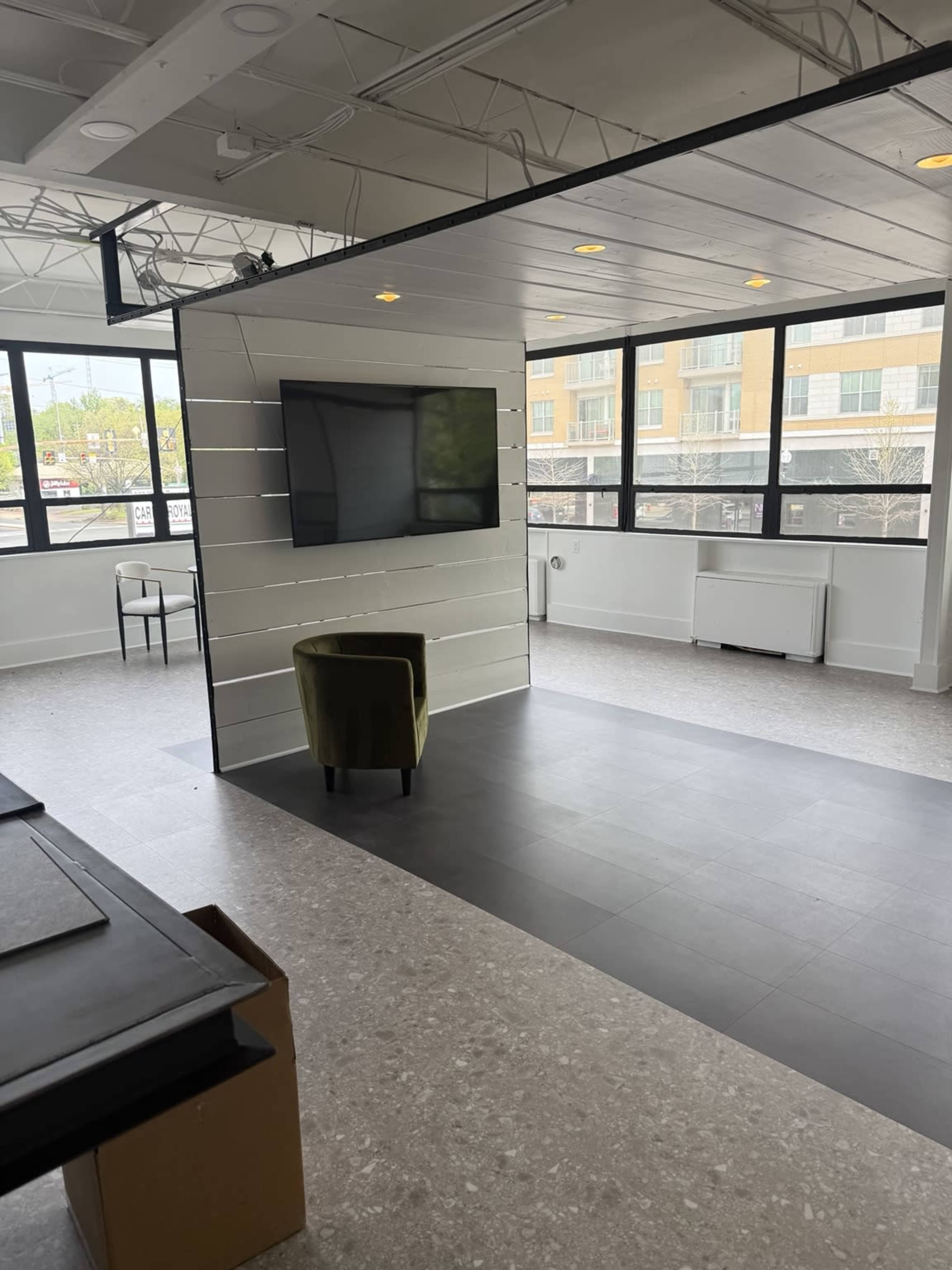 The image shows a modern office space with large windows, a wall-mounted TV, and a green chair positioned nearby.